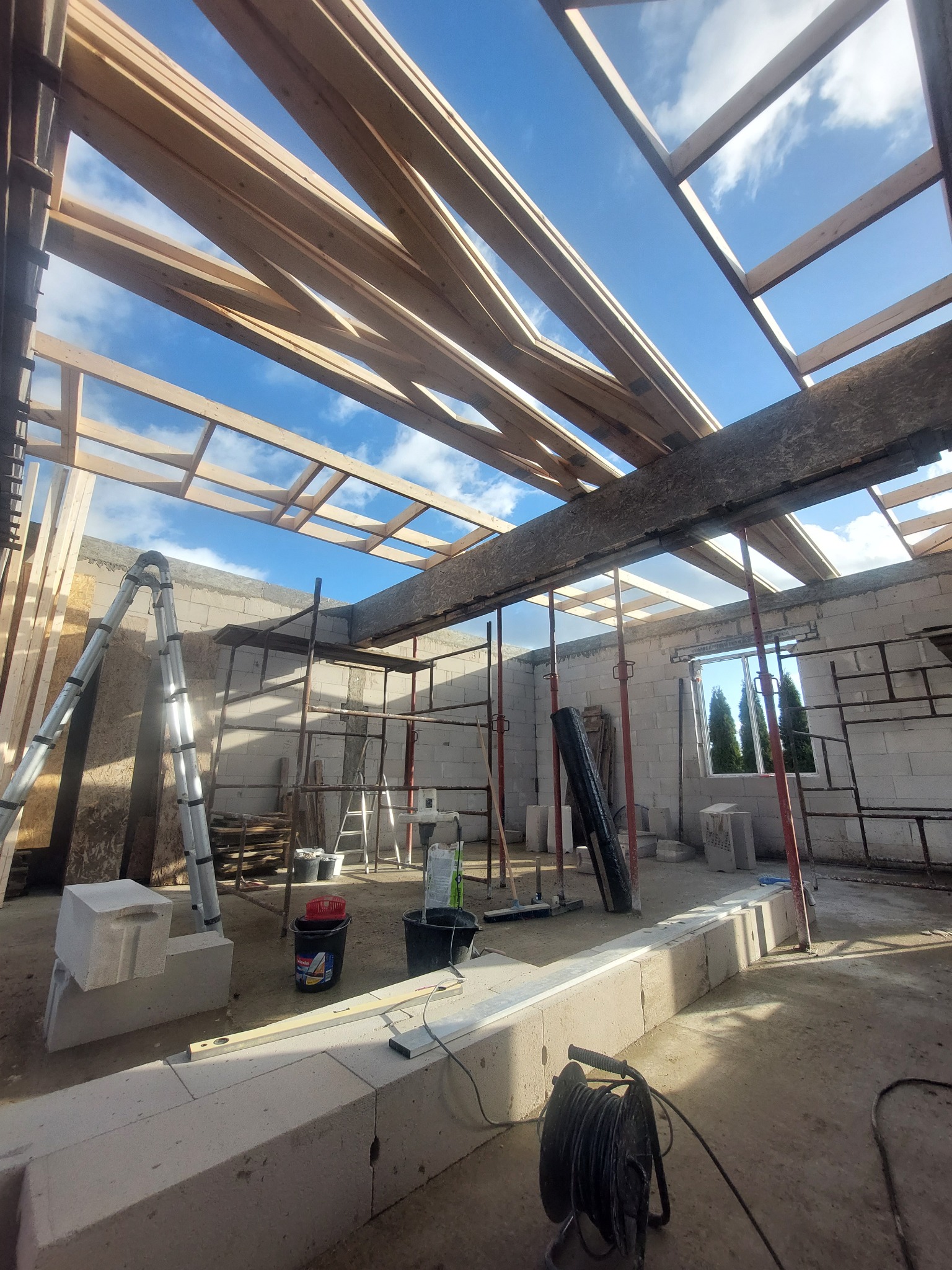 Wnętrze budynku w trakcie budowy, widoczne ściany z betonu komórkowego, drewniana konstrukcja dachu, rusztowania, drabina i narzędzia budowlane w nieładzie.