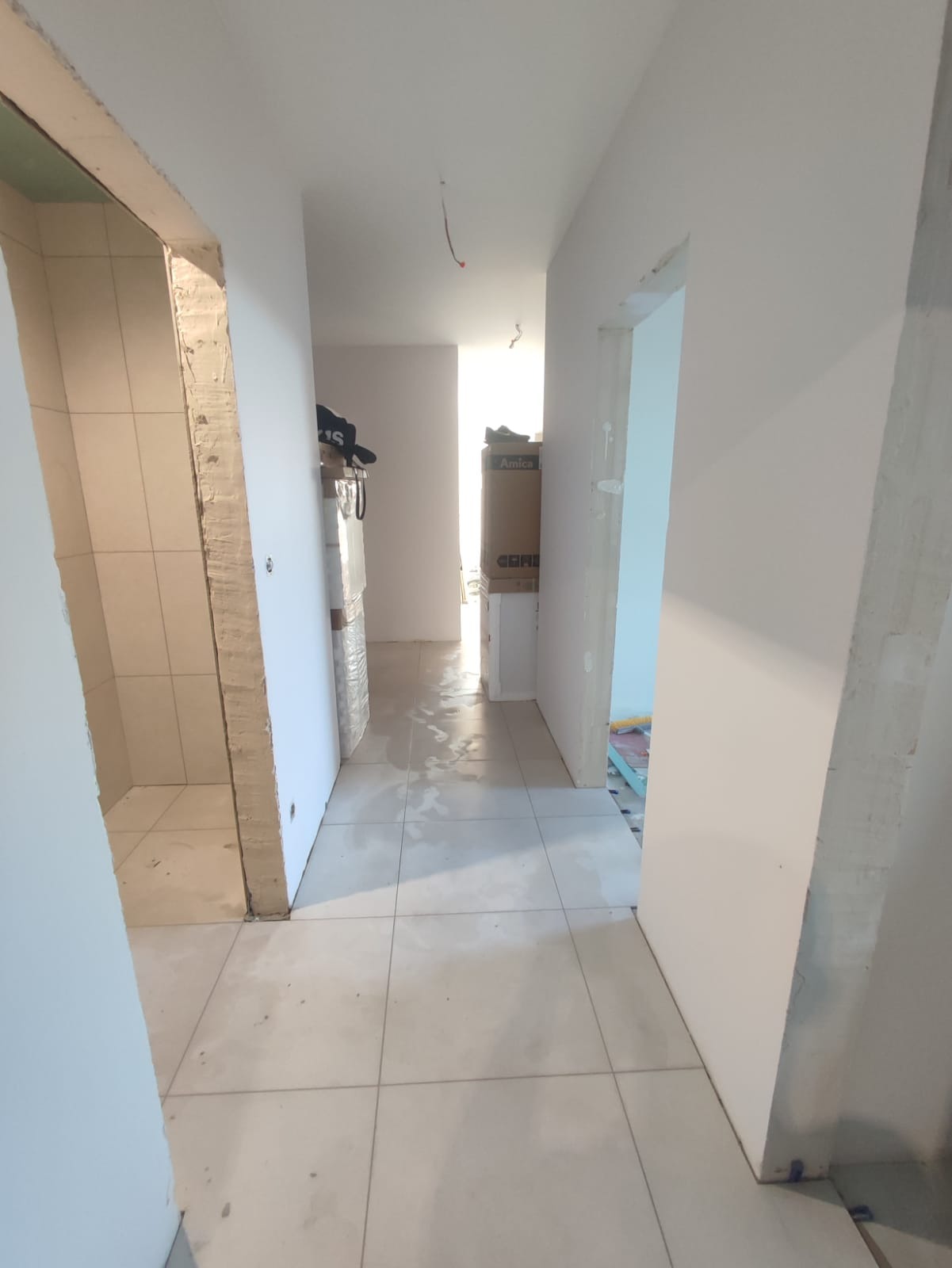 Korytarz w trakcie prac wykończeniowych: położona jasna, kwadratowa glazura podłogowa, widoczne surowe ościeżnice drzwiowe, ściany przygotowane pod malowanie, w głębi widoczne kartony...