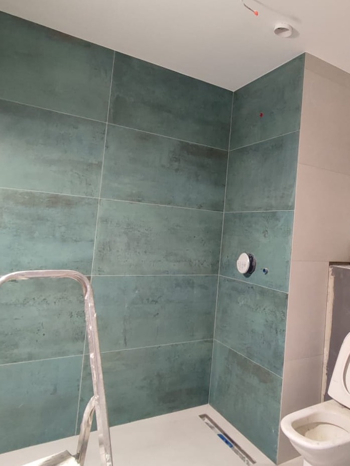 Wnętrze łazienki w trakcie prac wykończeniowych: poziome, turkusowe płytki imitujące metal na ścianach kabiny prysznicowej, widoczny odpływ liniowy, drabina malarska i częściowo zamontowana toaleta.