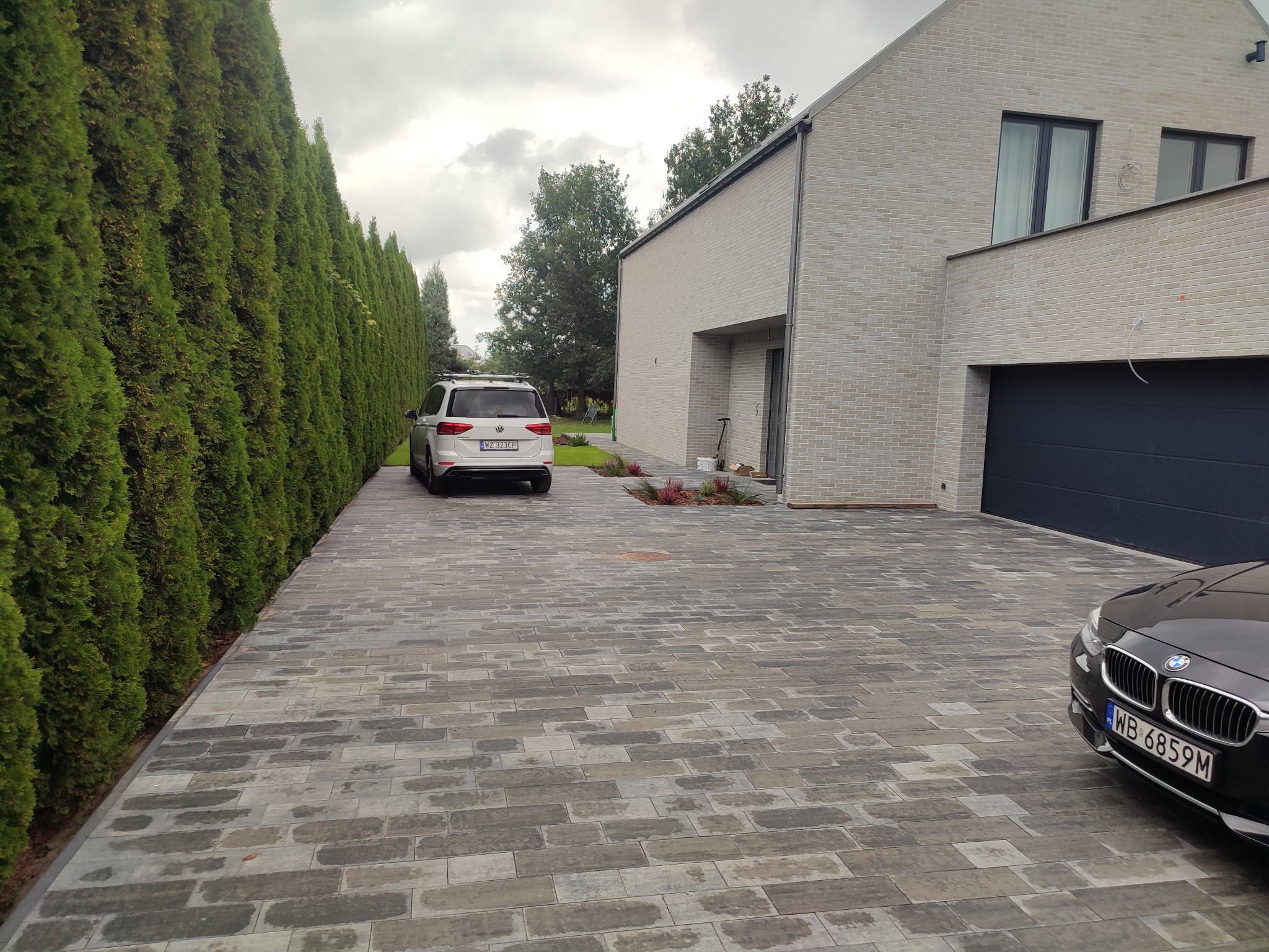 Nowoczesny podjazd z szarej kostki brukowej ułożonej w prostokątnym wzorze, obok żywopłotu z tui i jasnego domu z garażem.