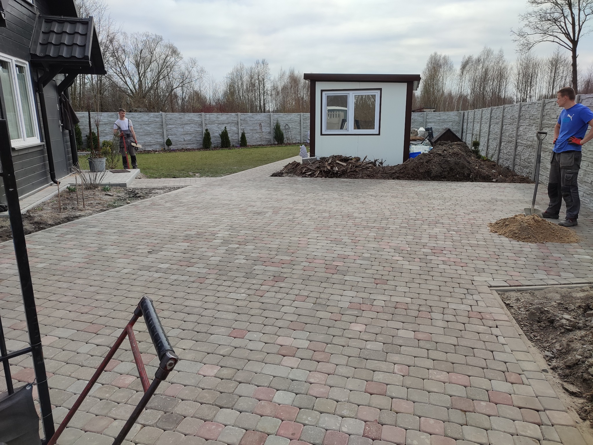 Rozległy podjazd z kostki w odcieniach szarości i czerwieni, częściowo ukończony, z widocznymi pracownikami i stertą ziemi w tle, ogrodzenie betonowe oraz budynek gospodarczy.
