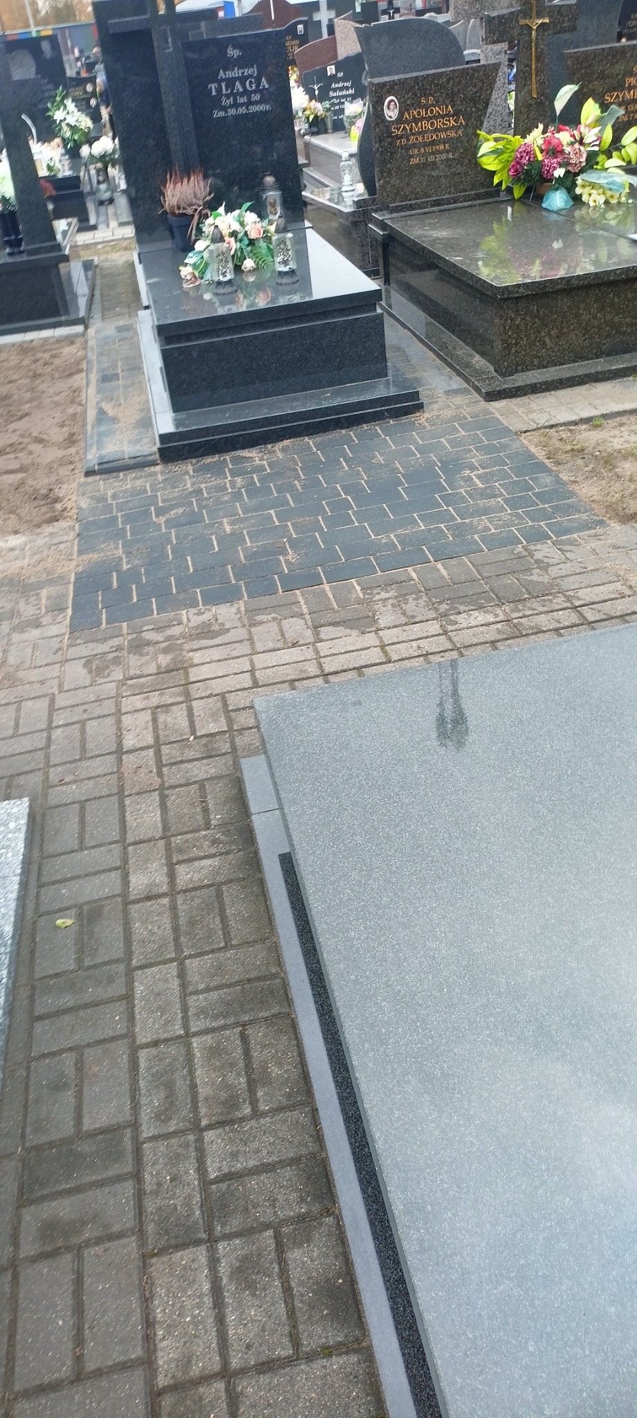 Układanie kostki brukowej w Stanisławowie wokół nagrobków, z widocznymi różnymi wzorami i kolorami kostki, prowadzące do pomników z kwiatami i zniczami.