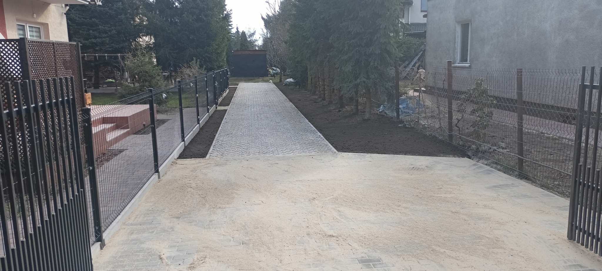 Ułożona szara kostka brukowa tworząca ścieżkę i plac przed domem, obrzeża wykończone czarną ziemią, ogrodzenie z siatki po obu stronach, w tle rząd drzew iglastych i budynek.