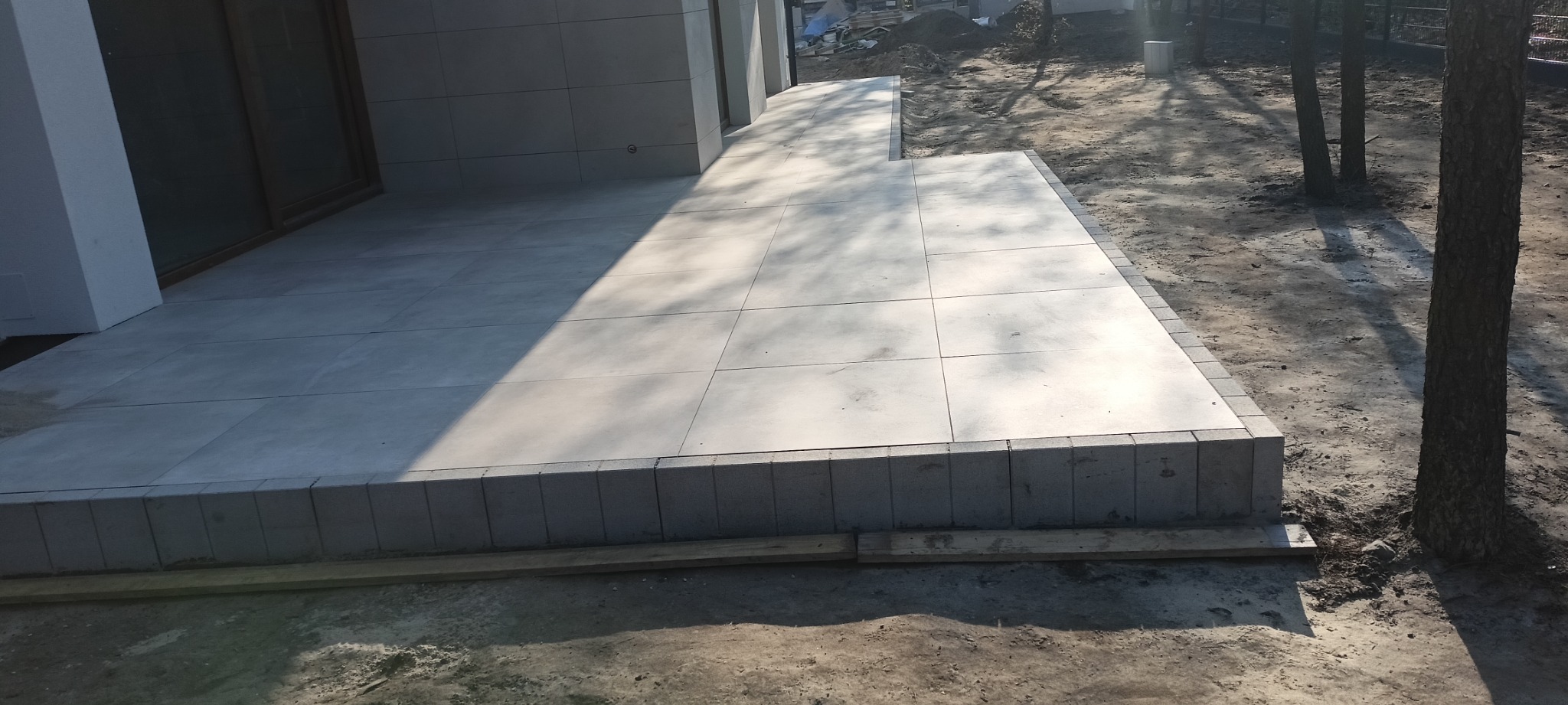 Nowoczesny taras z jasnych, kwadratowych płyt, obrzeża wykończone drobną kostką, widoczne drewniane deski pod konstrukcją, w tle ziemia i drzewa.