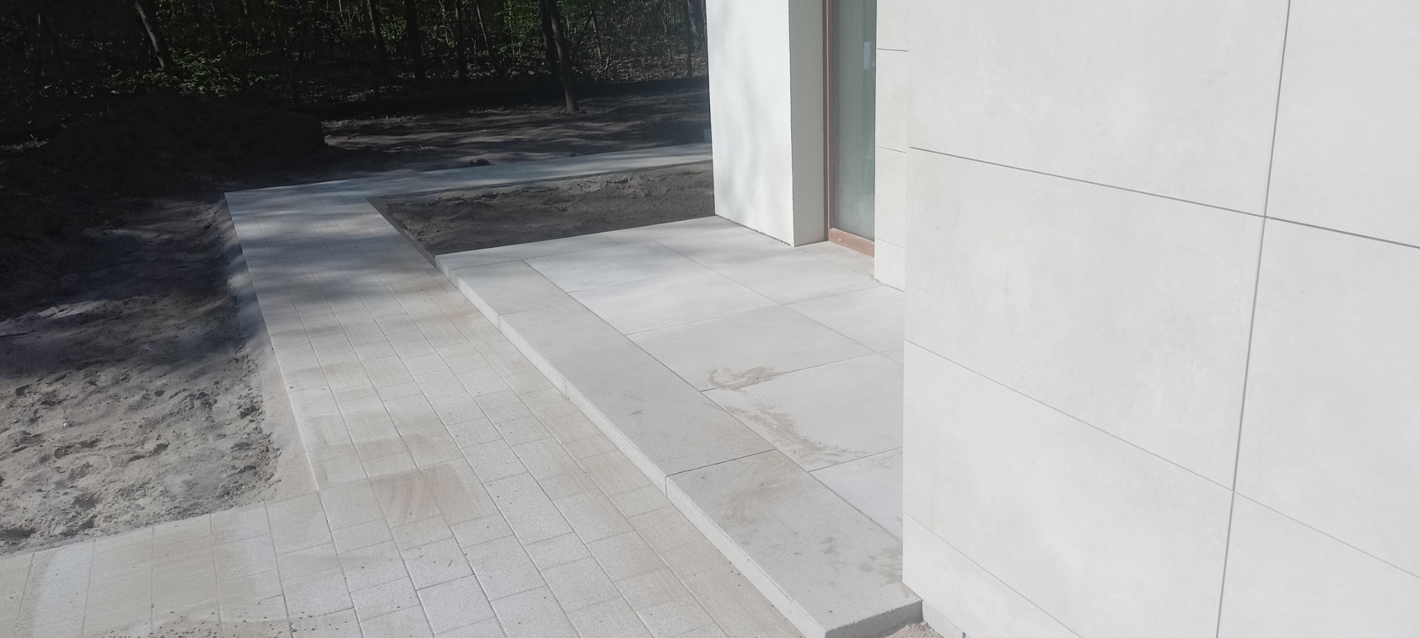 Jasnoszara kostka brukowa prowadząca do wejścia domu z jasnymi płytami elewacyjnymi, widoczny fragment rabaty ziemnej z tyłu.