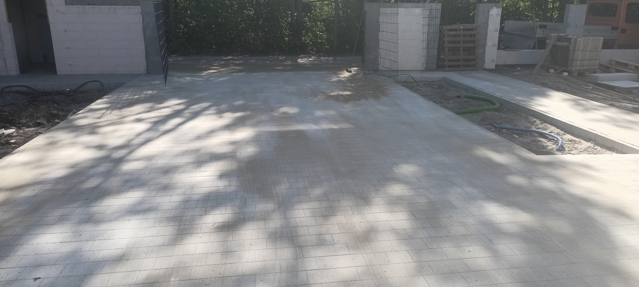 Nowa, jasnoszara nawierzchnia z kostki brukowej w trakcie budowy, widoczne elementy konstrukcyjne budynku w tle i cień drzew na powierzchni.