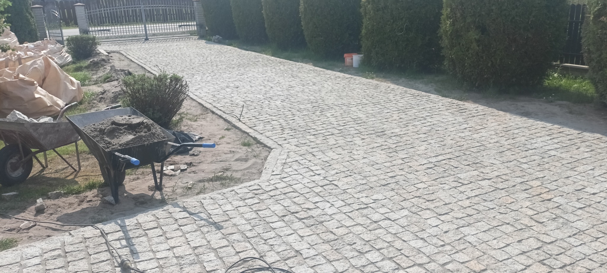 Ułożona granitowa kostka brukowa na podjeździe, widoczne dwie taczki: jedna z piaskiem i druga z kamieniami, w tle rząd tuj.