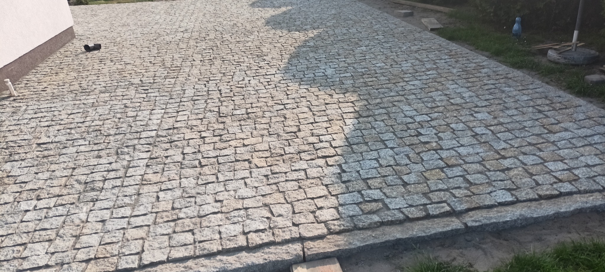 Ułożona nawierzchnia z kostki granitowej w odcieniach szarości, widoczna z góry, z elementami otoczenia jak fragment budynku i trawnik.
