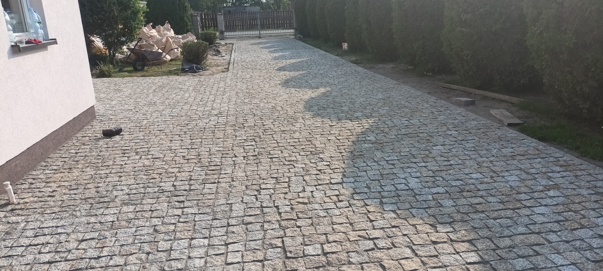 Ułożona z kostki granitowej ścieżka prowadząca do bramy, z workami i taczką w tle, widok z perspektywy chodnika.