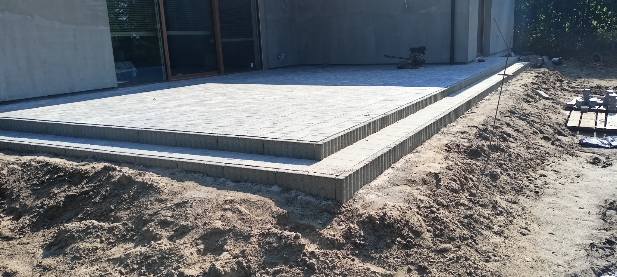 Nowy taras z kostki brukowej z dwoma stopniami, widoczne nierówności terenu wokół i zagęszczarka spalinowa w tle.