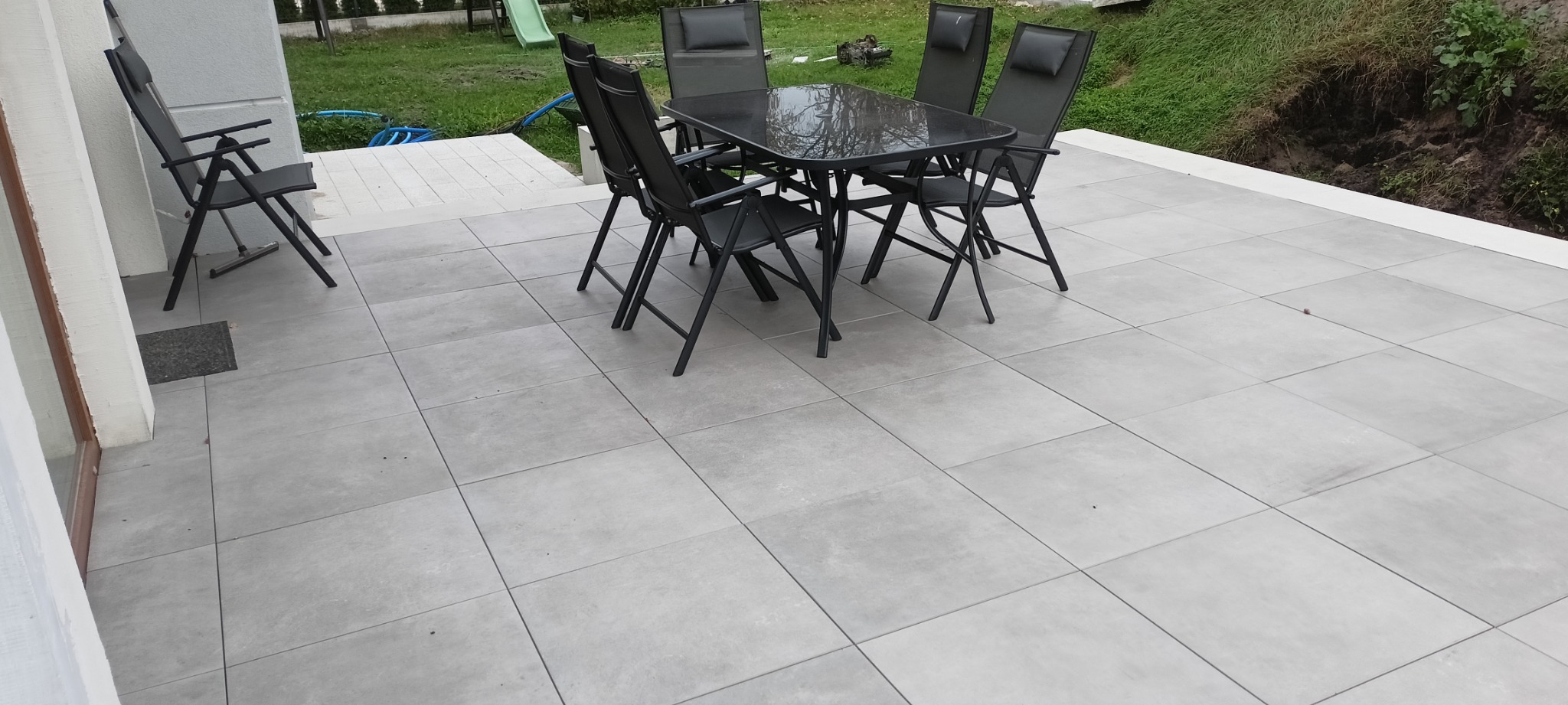 Jasnoszara, kwadratowa nawierzchnia tarasu z kompletem mebli ogrodowych: szklany stół i czarne krzesła z oparciami. Widoczny fragment budynku z białym wykończeniem i trawnik w tle.