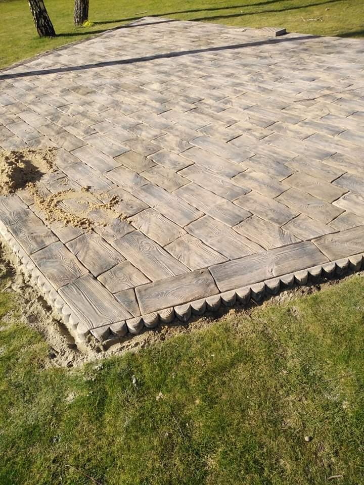 Ułożona nawierzchnia z kostki imitującej drewno, z dekoracyjnym obrzeżem, częściowo zasypana piaskiem, widoczne trawnik i drzewa w tle.