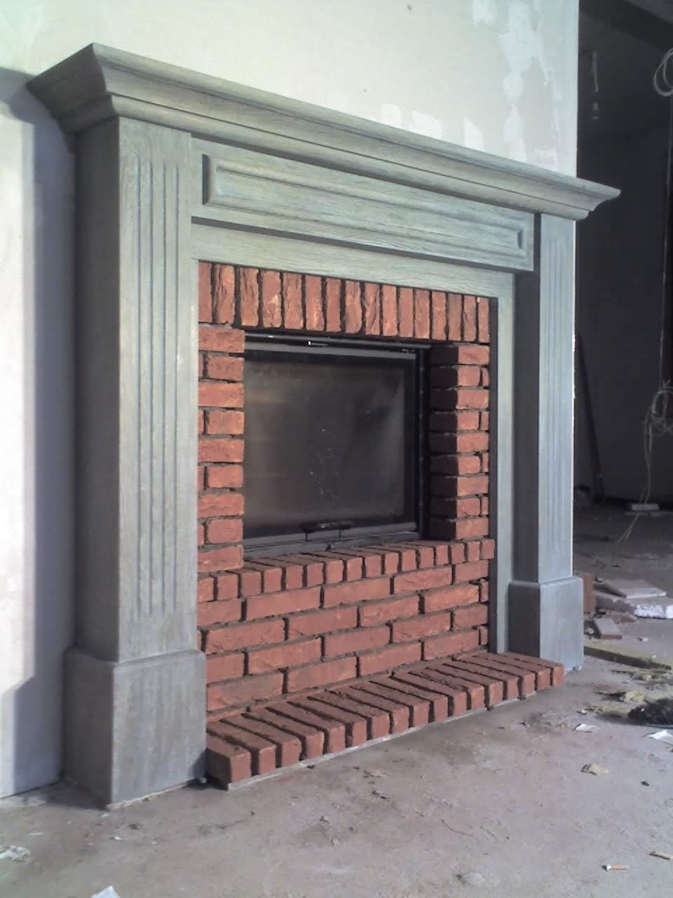 Niedokończony kominek z cegły z szarym obramowaniem w surowym wnętrzu, widoczne okablowanie i resztki materiałów budowlanych.
