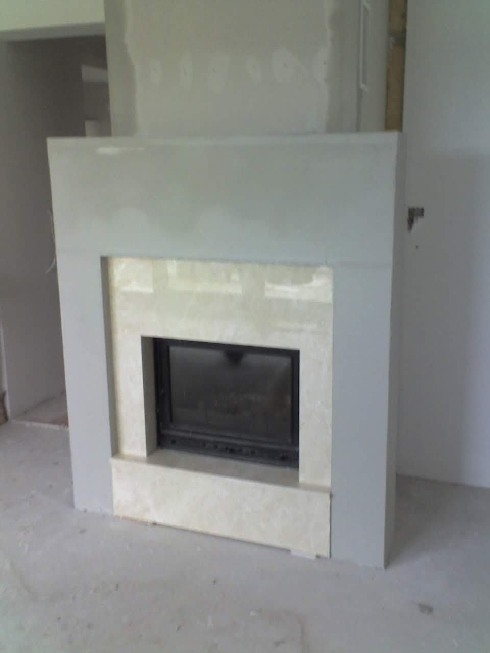Niedokończony kominek z marmurowym obramowaniem i szarym tynkiem, widoczne ślady prac wykończeniowych na ścianach i podłodze.