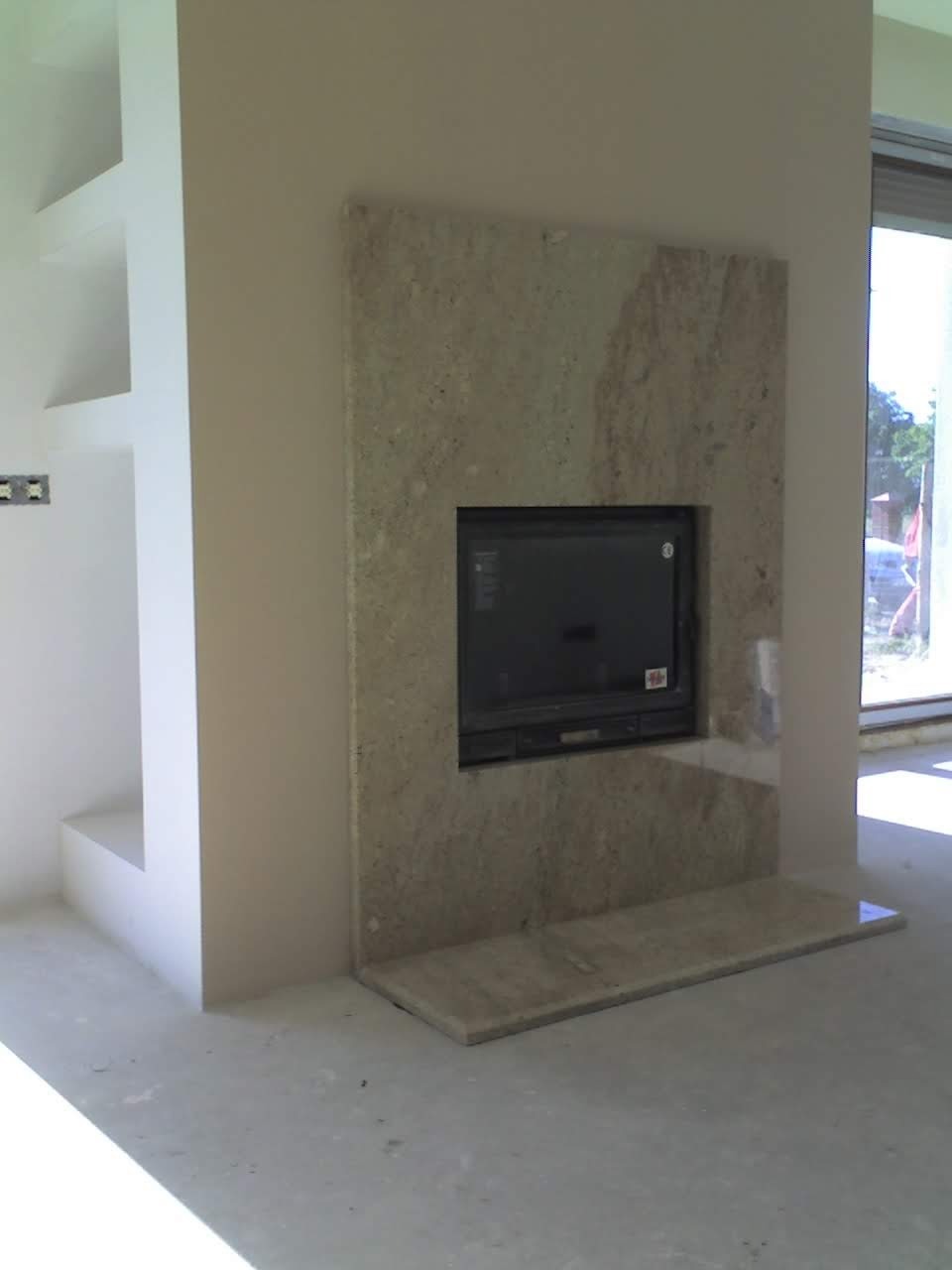 Obudowa kominkowa z jasnego kamienia z ciemnym wkładem, w surowym wnętrzu z widoczną wnęką w ścianie i dużym oknem w tle.