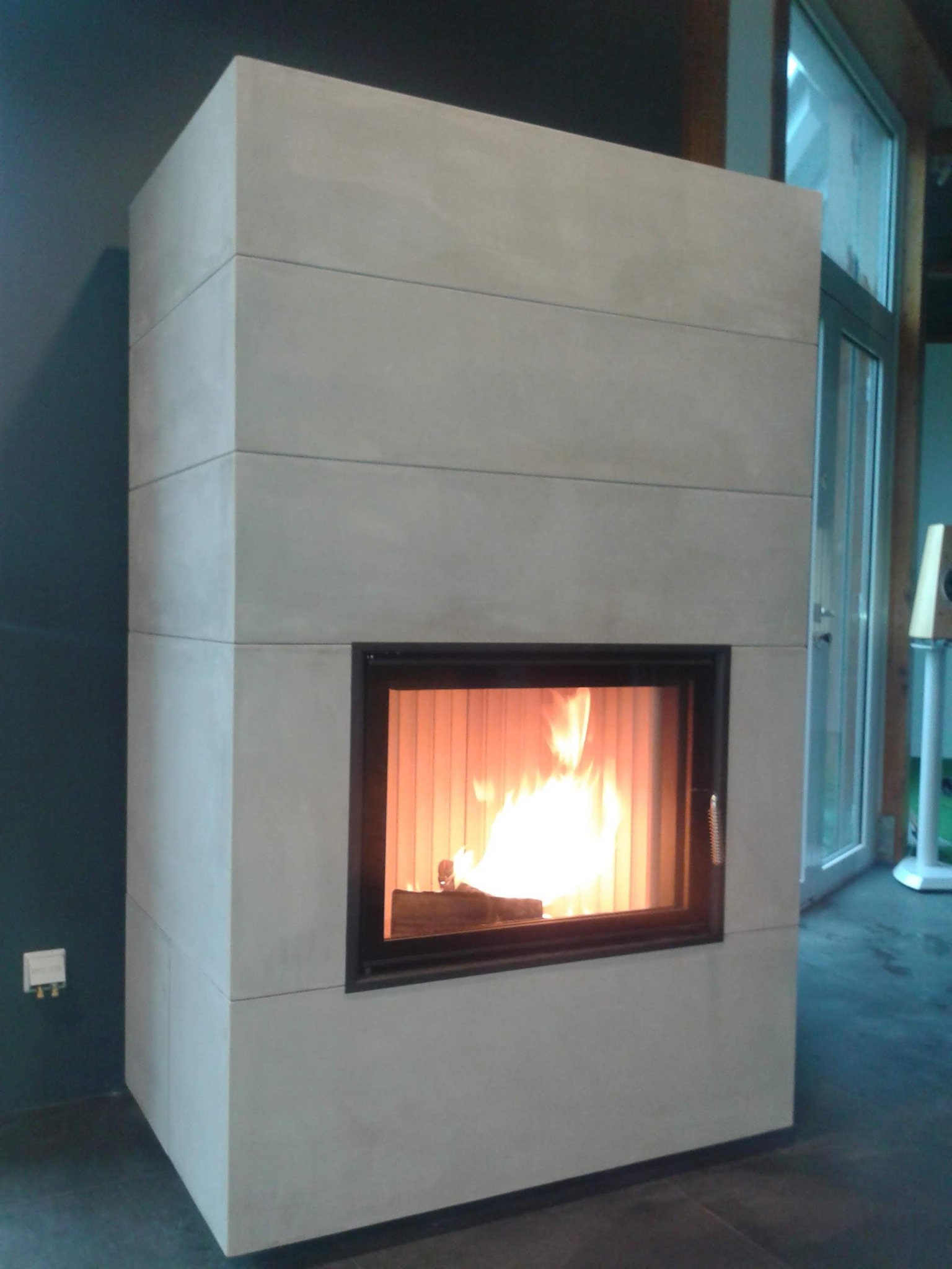 Nowoczesny kominek obudowany jasnoszarymi płytami, z widocznym płomieniem i czarną ramą paleniska, ustawiony w pomieszczeniu z ciemną ścianą i przeszklonymi drzwiami balkonowymi.
