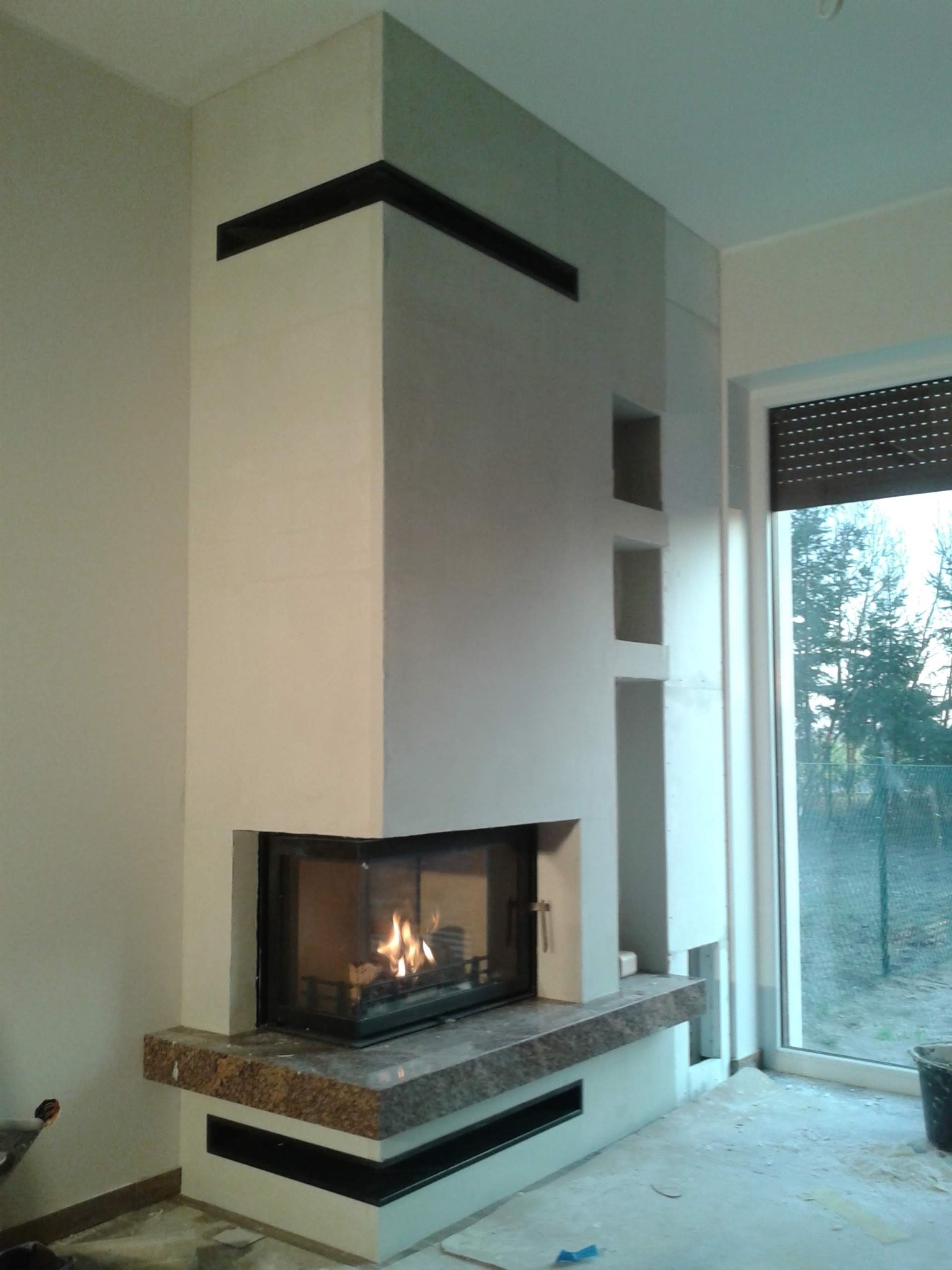 Nowoczesny kominek narożny z paleniskiem, wykończony jasnym tynkiem strukturalnym i blatem z brązowego granitu, z wnękami dekoracyjnymi po prawej stronie, widok na zewnątrz przez okno z roletą.