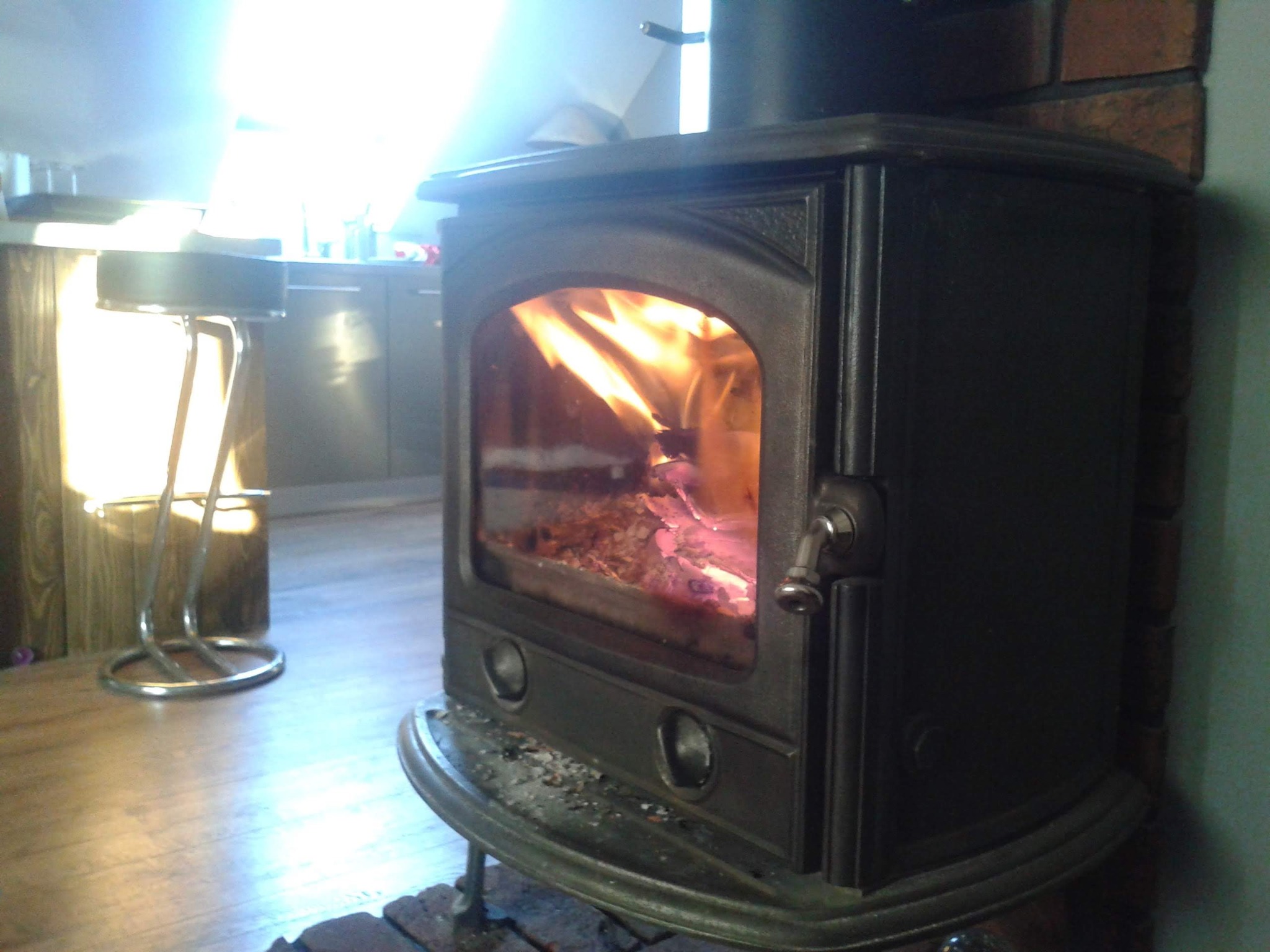 Czarny, żeliwny piec kominkowy z płomieniami widocznymi przez szybę, stojący w pomieszczeniu z drewnianą podłogą i fragmentem baru z chromowanym stołkiem.