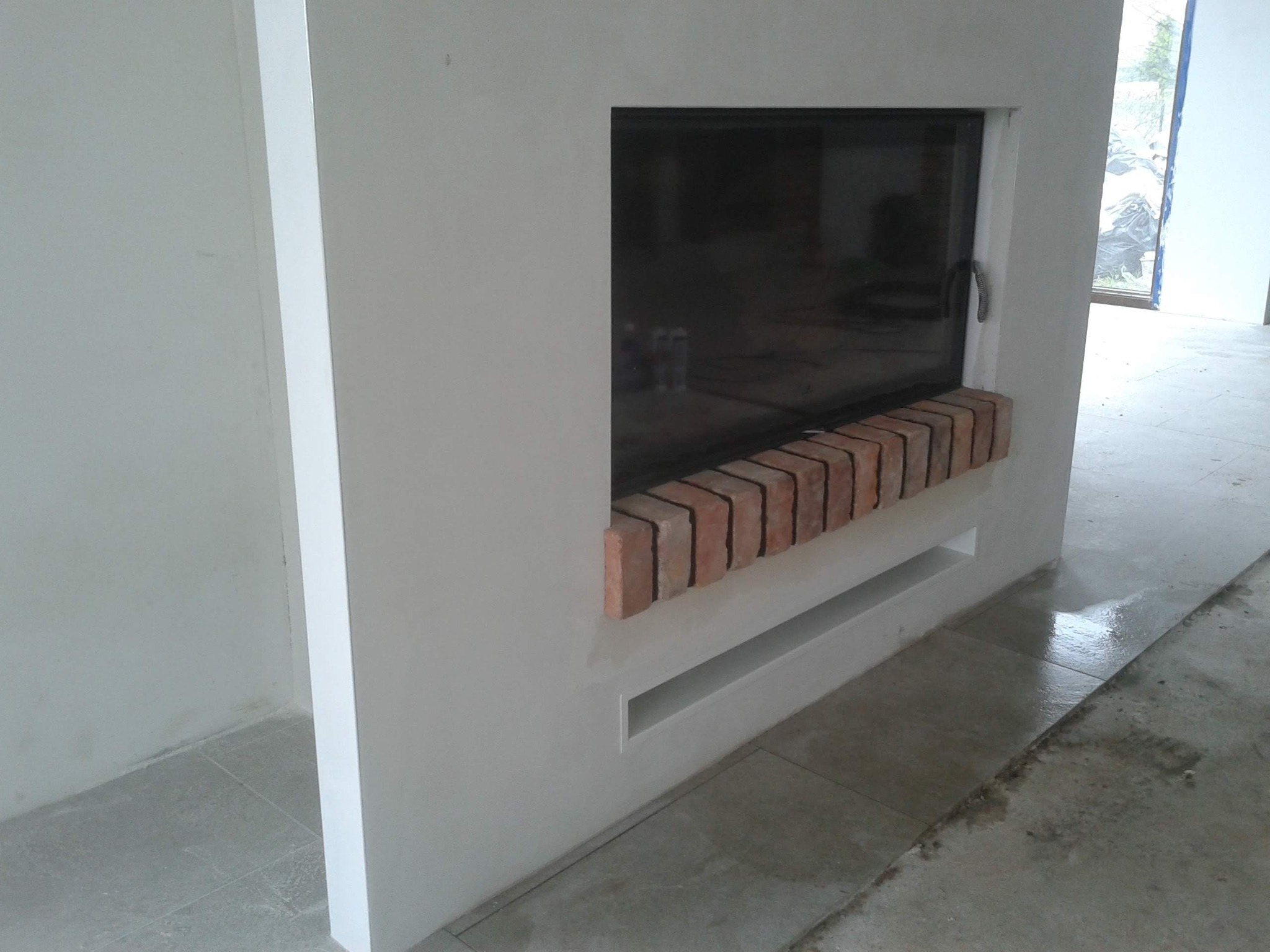 Wnęka kominkowa w trakcie wykańczania, z ceglanym obramowaniem i widocznym wkładem kominkowym, surowy stan ścian i podłogi.
