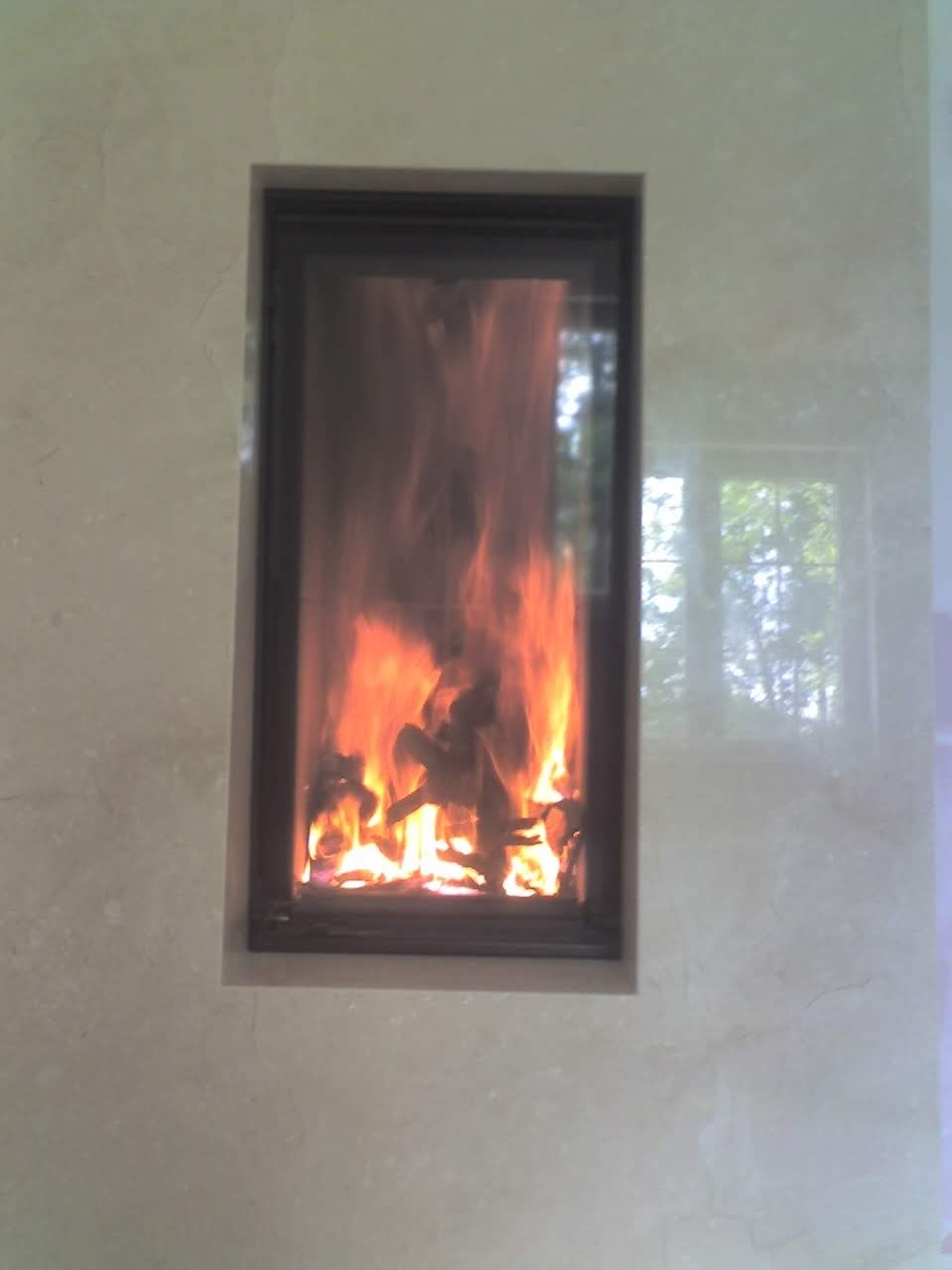 Wbudowany kominek z czarną ramą i palącym się drewnem, otoczony jasnym marmurem z widocznym odbiciem okna.