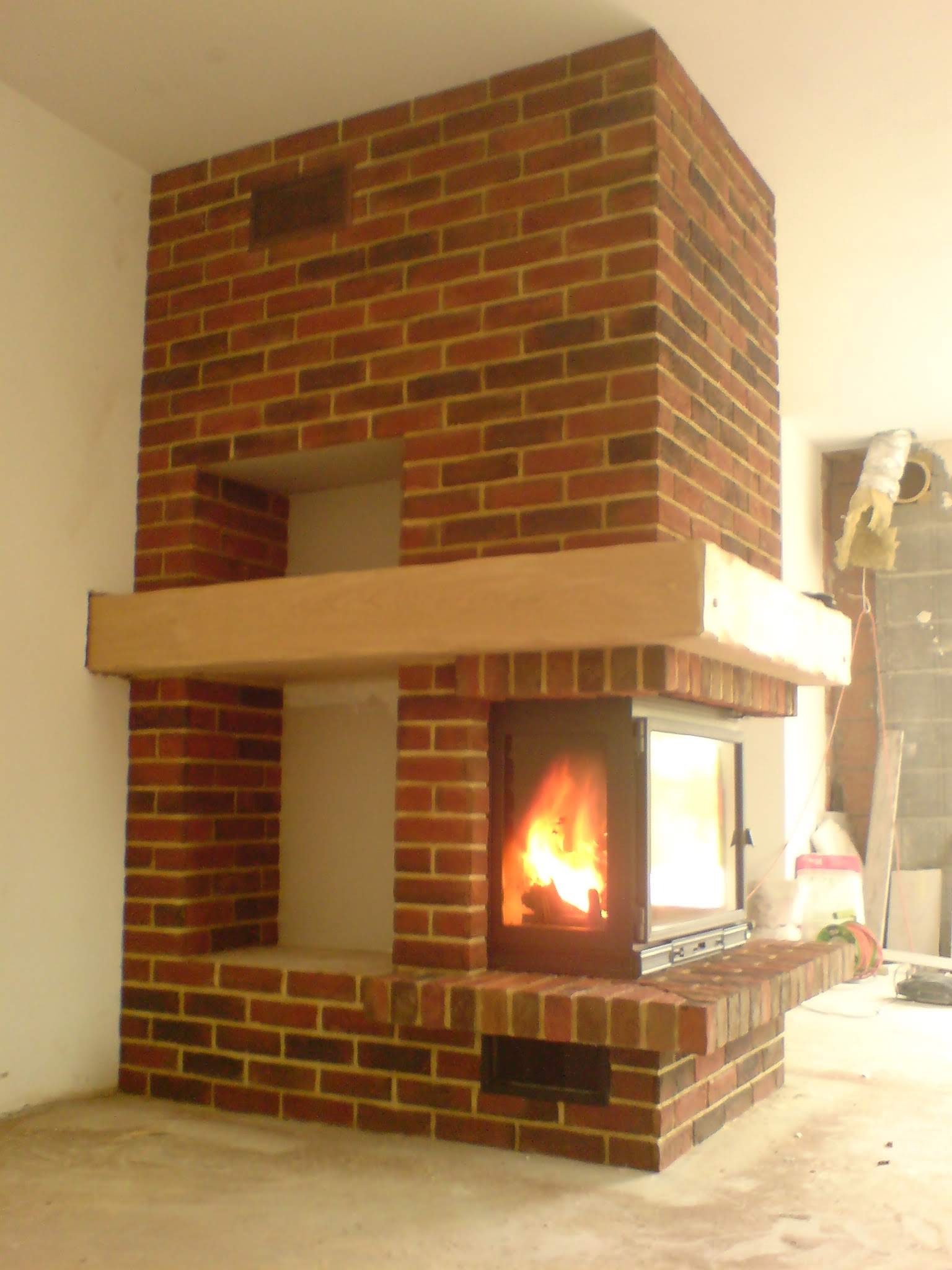 Murowany kominek z czerwonej cegły z drewnianą półką, wkład kominkowy z palącym się ogniem, surowy stan wykończenia wnętrza.