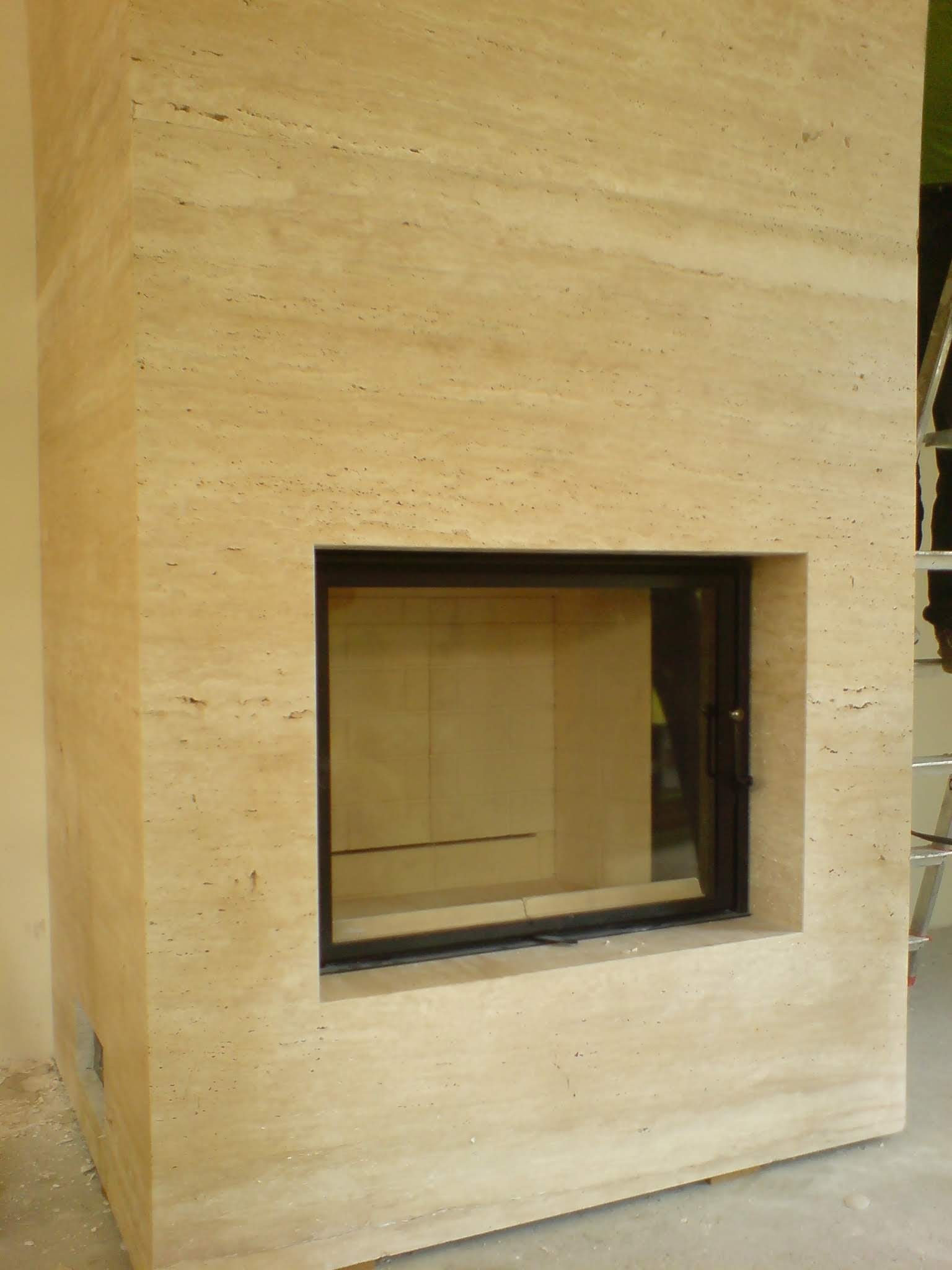 Obudowa kominka z trawertynu z czarną ramą, widoczne niedokończone wykończenie ściany i drabina w tle.