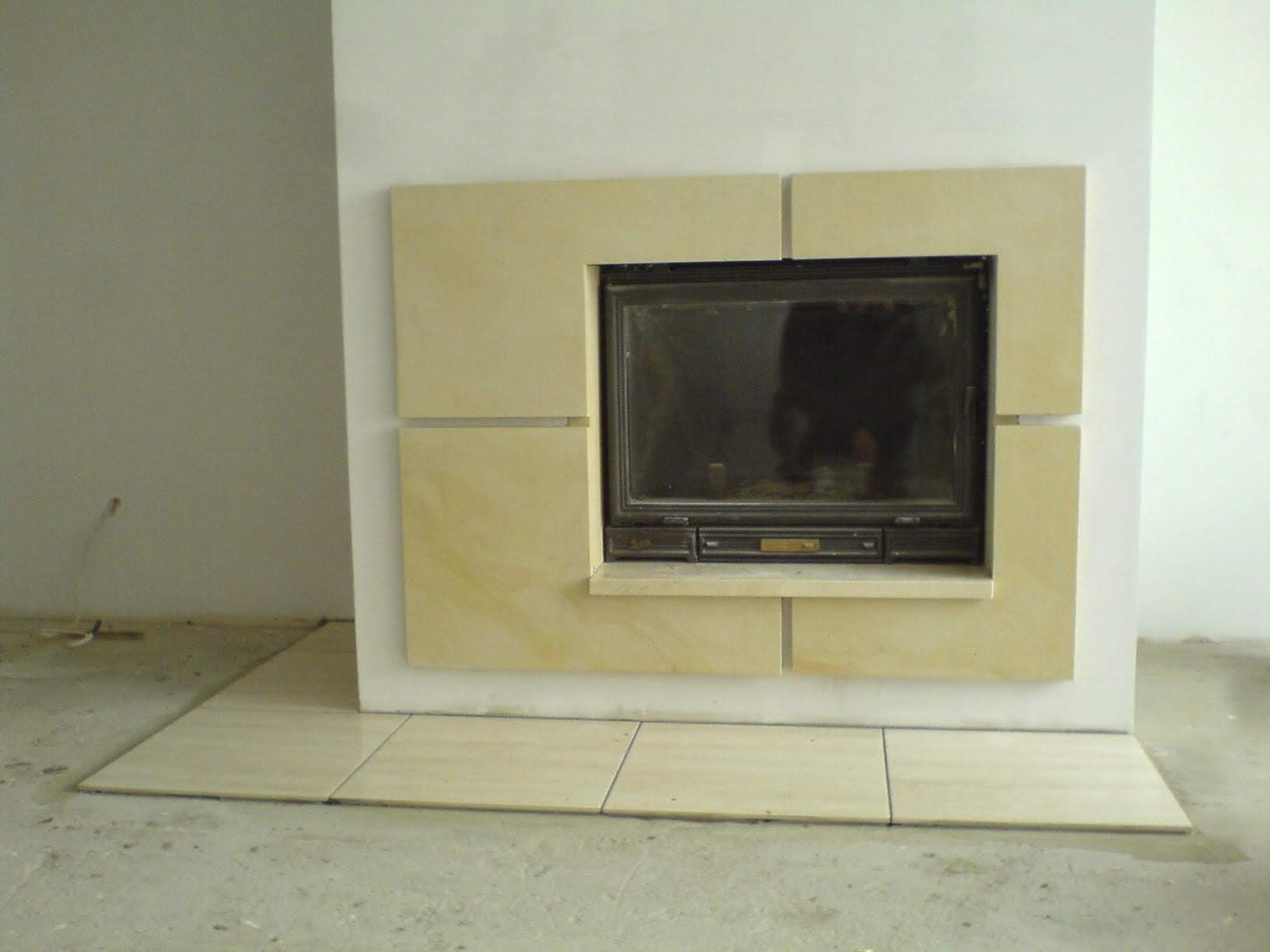 Wkład kominkowy z obudową z jasnego kamienia, widoczne płytki podłogowe w kolorze obudowy, surowy stan wykończenia wnętrza.