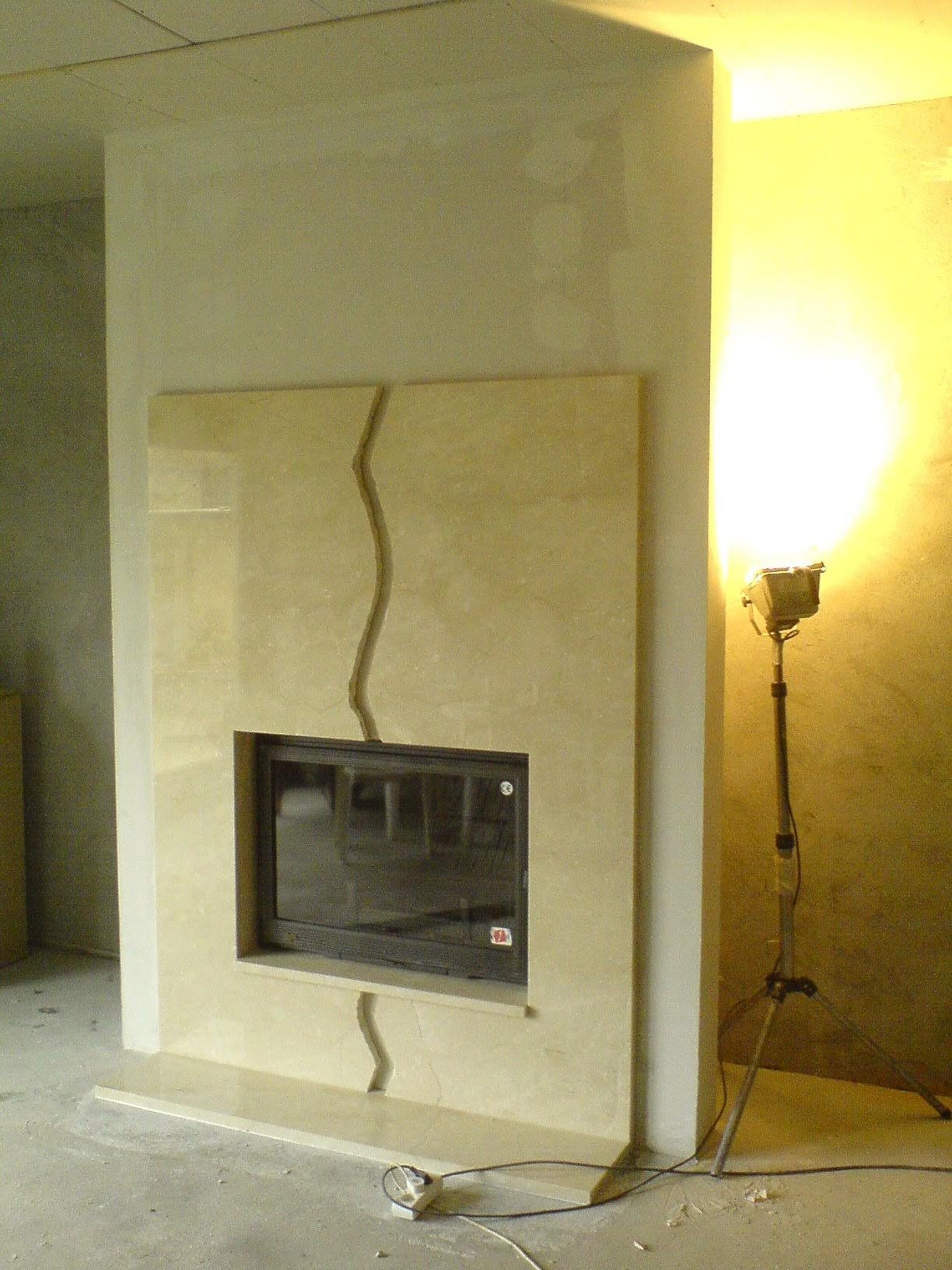 Beżowy kominek z czarnym wkładem, wykończony płytami z dekoracyjnym, falistym łączeniem, oświetlony lampą budowlaną na trójnogu w surowym wnętrzu.