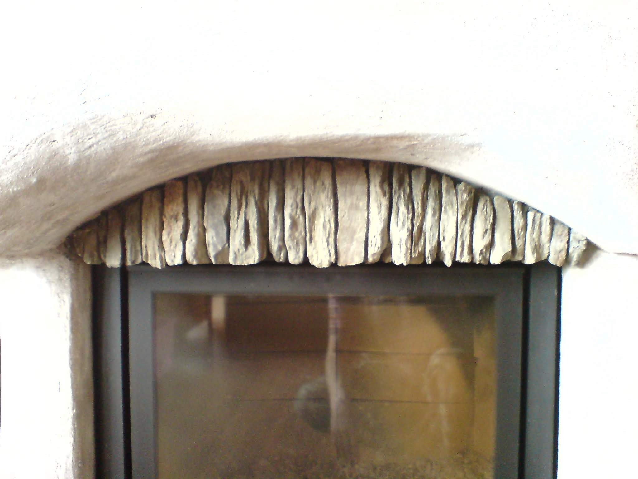 Fragment obudowy kominka z dekoracyjnym łukiem z kamiennych płytek nad szybą paleniska, biały tynk wokół.