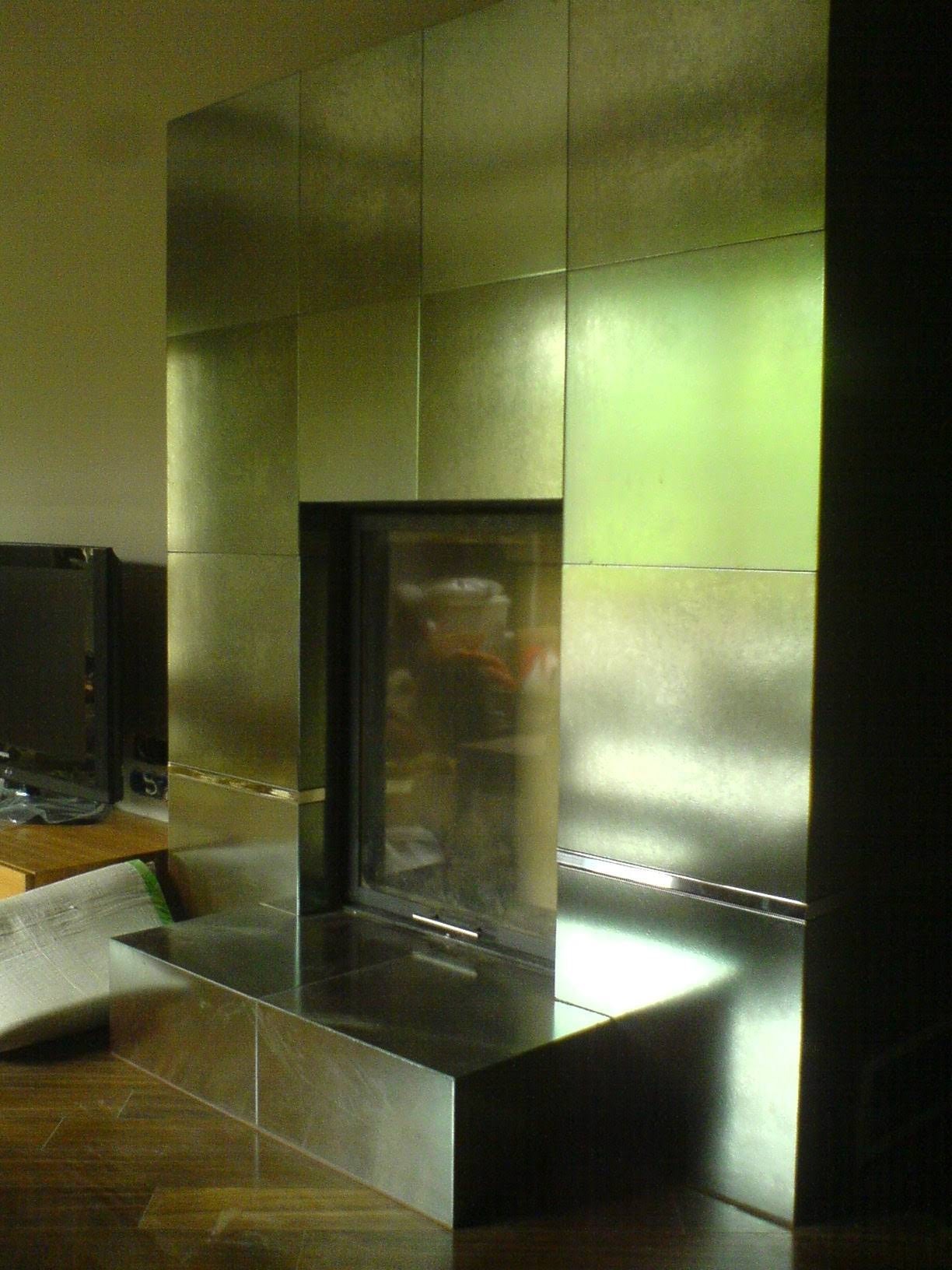 Nowoczesny kominek obłożony metalowymi płytami, z widocznym odbiciem w szkle paleniska i fragmentem telewizora w tle.