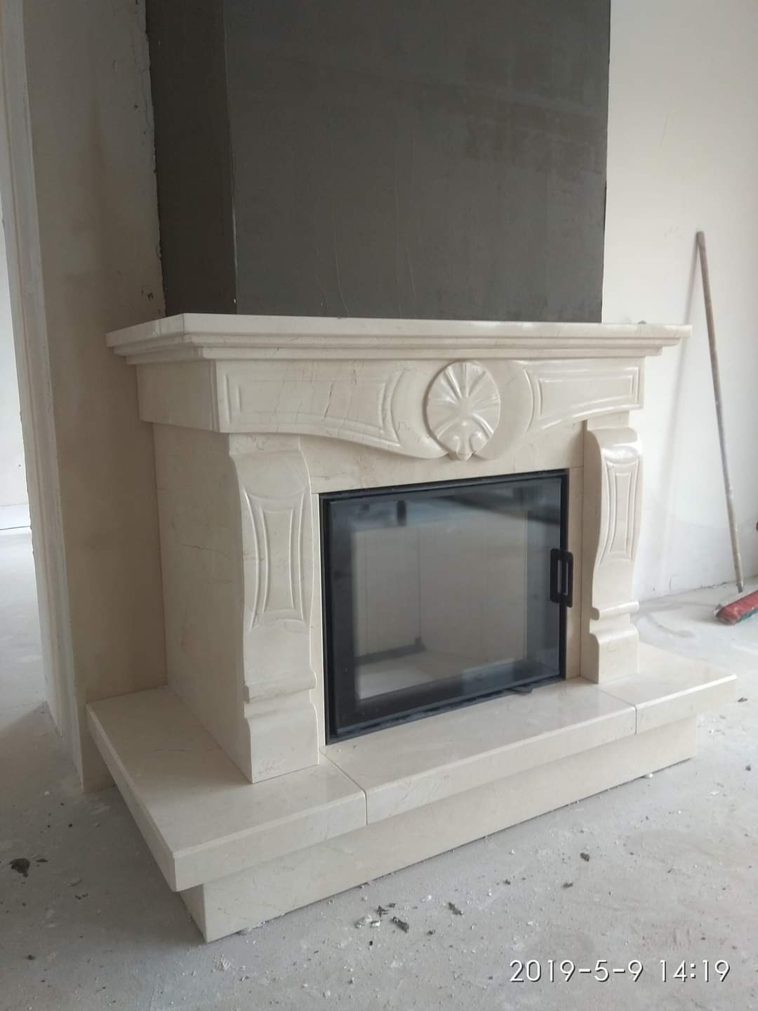 Marmurowy portal kominkowy z czarnymi drzwiczkami, widoczne niedokończone prace wykończeniowe na ścianie i podłodze.