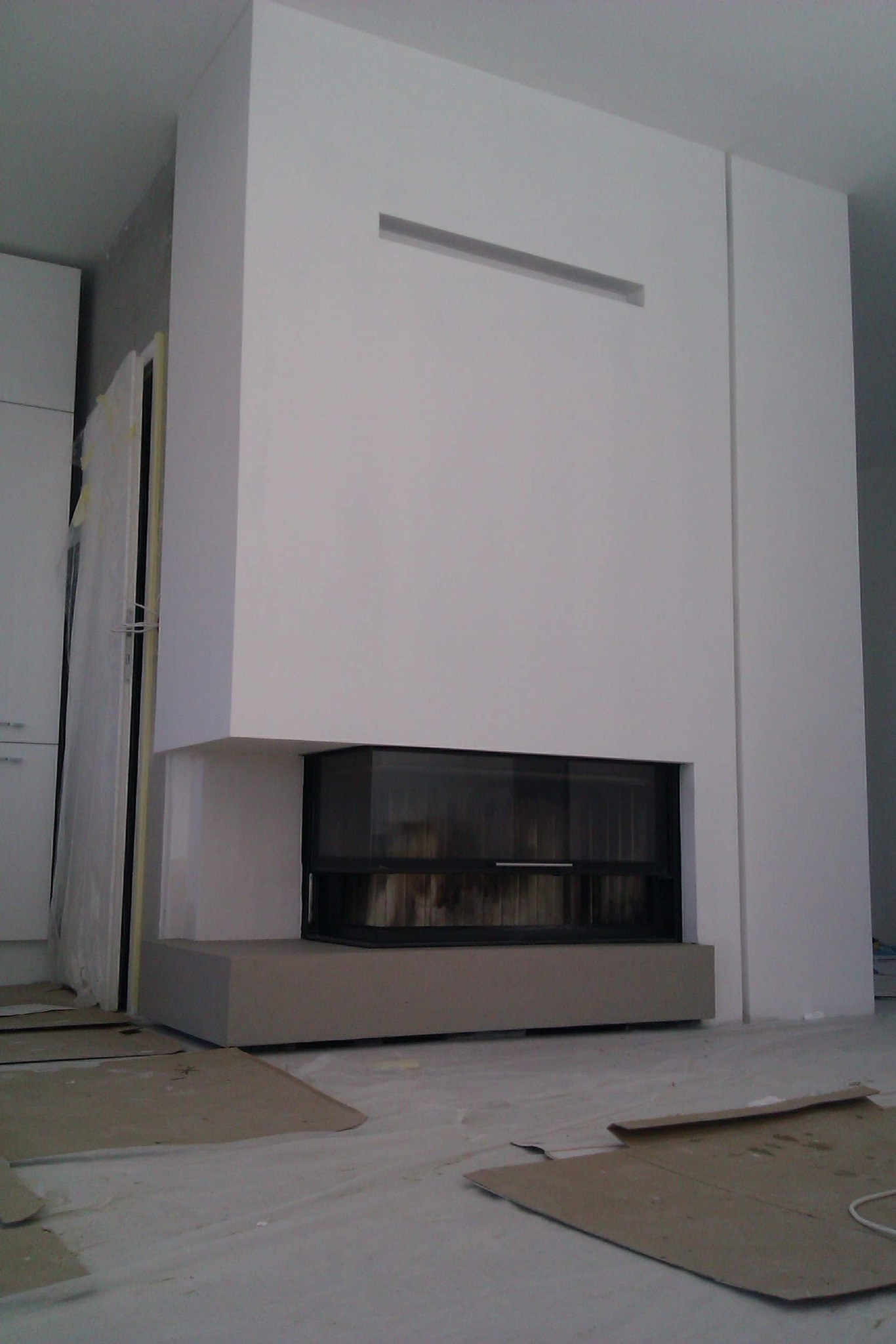 Nowoczesny kominek z prostokątnym wlotem powietrza w białej obudowie, widoczne zabezpieczenia podłogi kartonem i folią malarską podczas montażu.