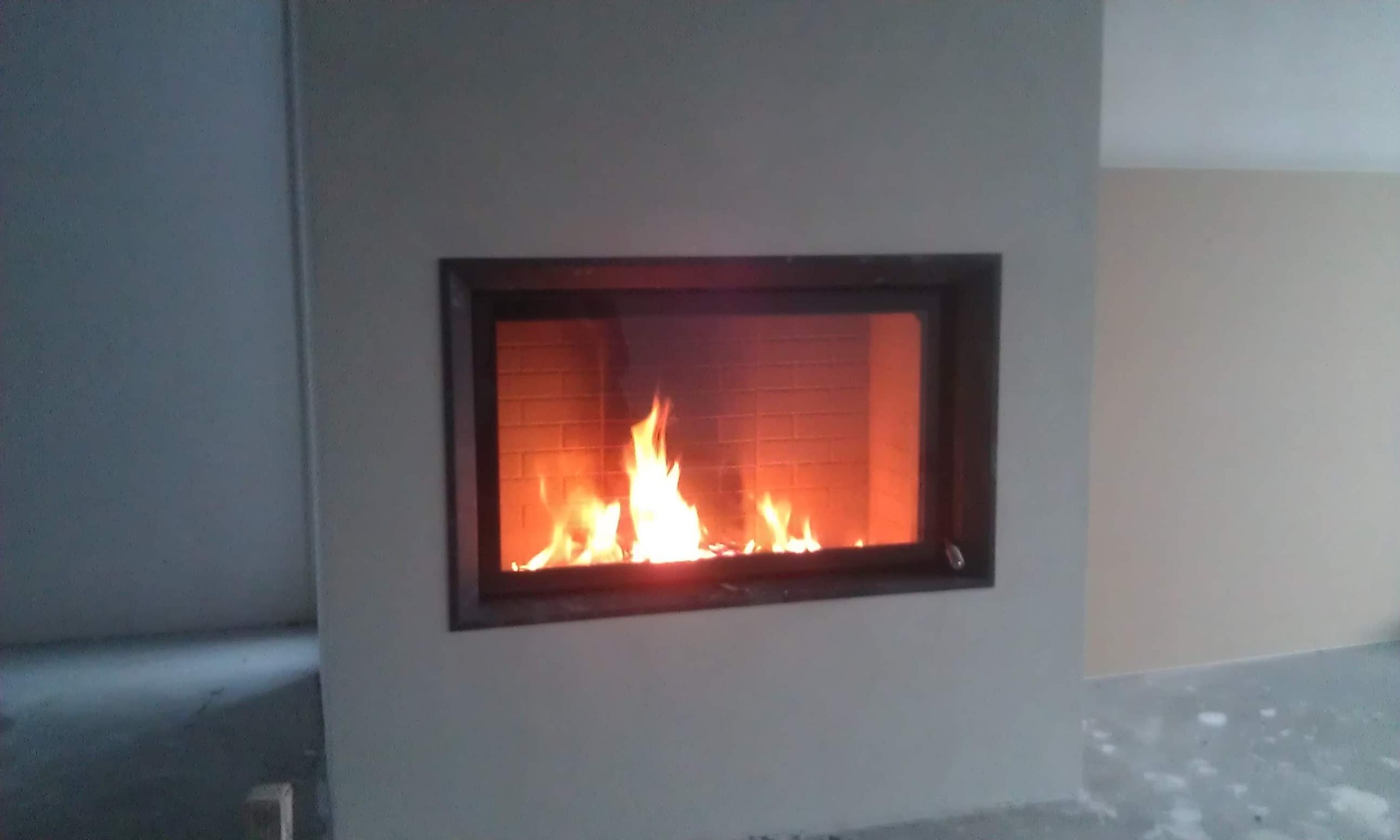 Nowoczesny kominek z szybą i ceglanym wnętrzem, płomienie tańczą w palenisku, surowe wykończenie ścian w odcieniach szarości i beżu. Widoczny fragment podłogi w trakcie prac wykończeniowych.