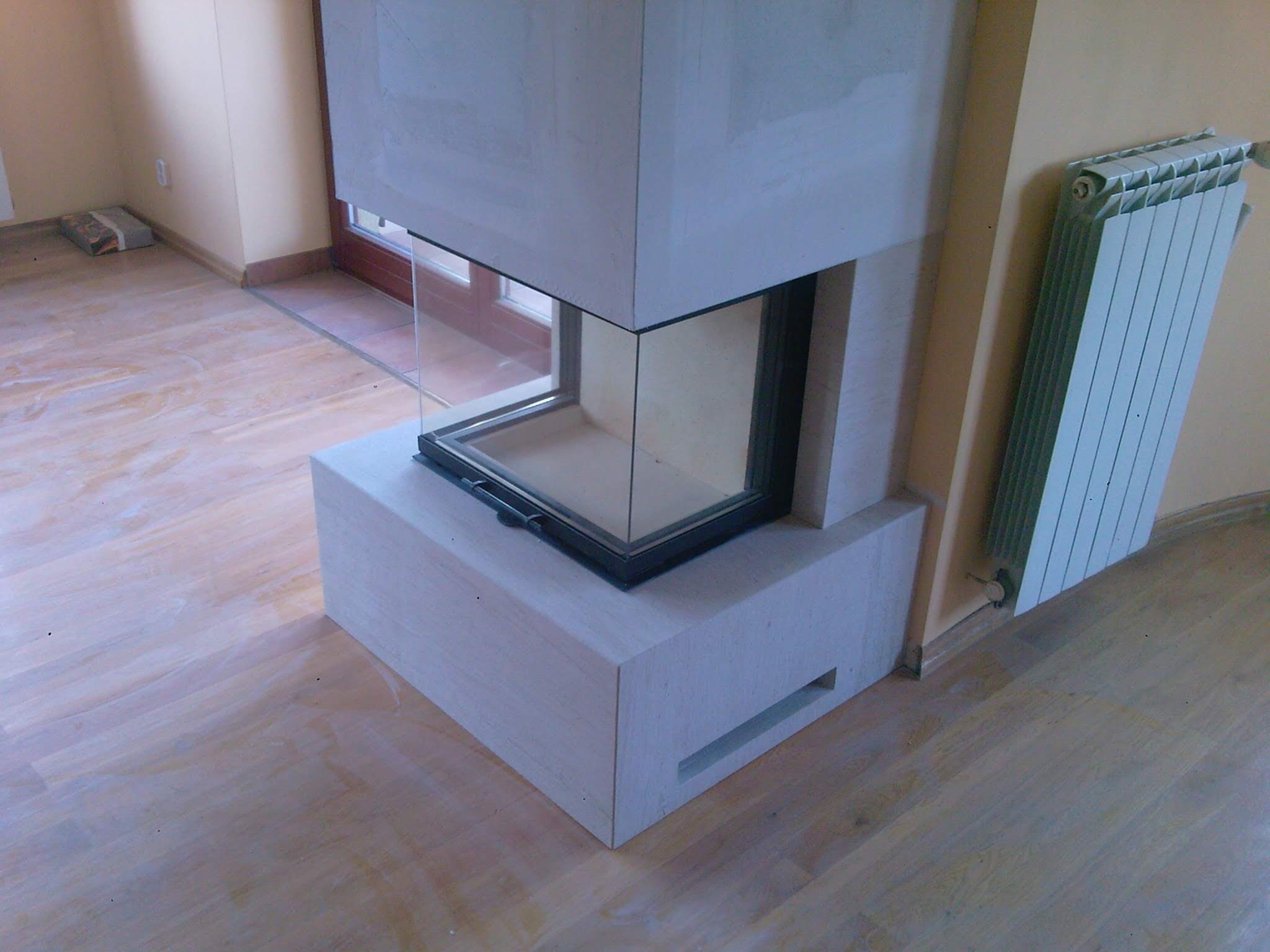 Narożny kominek z jasnego kamienia z szybą, zamontowany w salonie z drewnianą podłogą i grzejnikiem aluminiowym na ścianie.