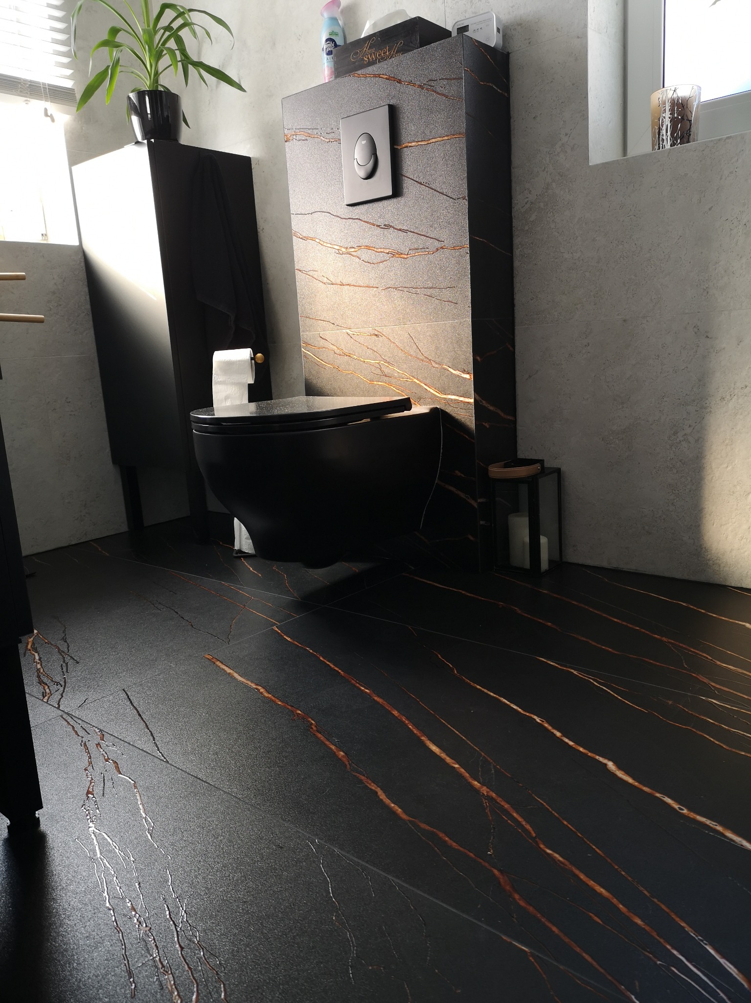 Nowoczesna łazienka z czarną ceramiką i płytkami imitującymi marmur z brązowymi żyłami, wisząca toaleta z zabudowanym systemem spłukiwania, minimalistyczna szafka umywalkowa i dekoracyjna roślina...