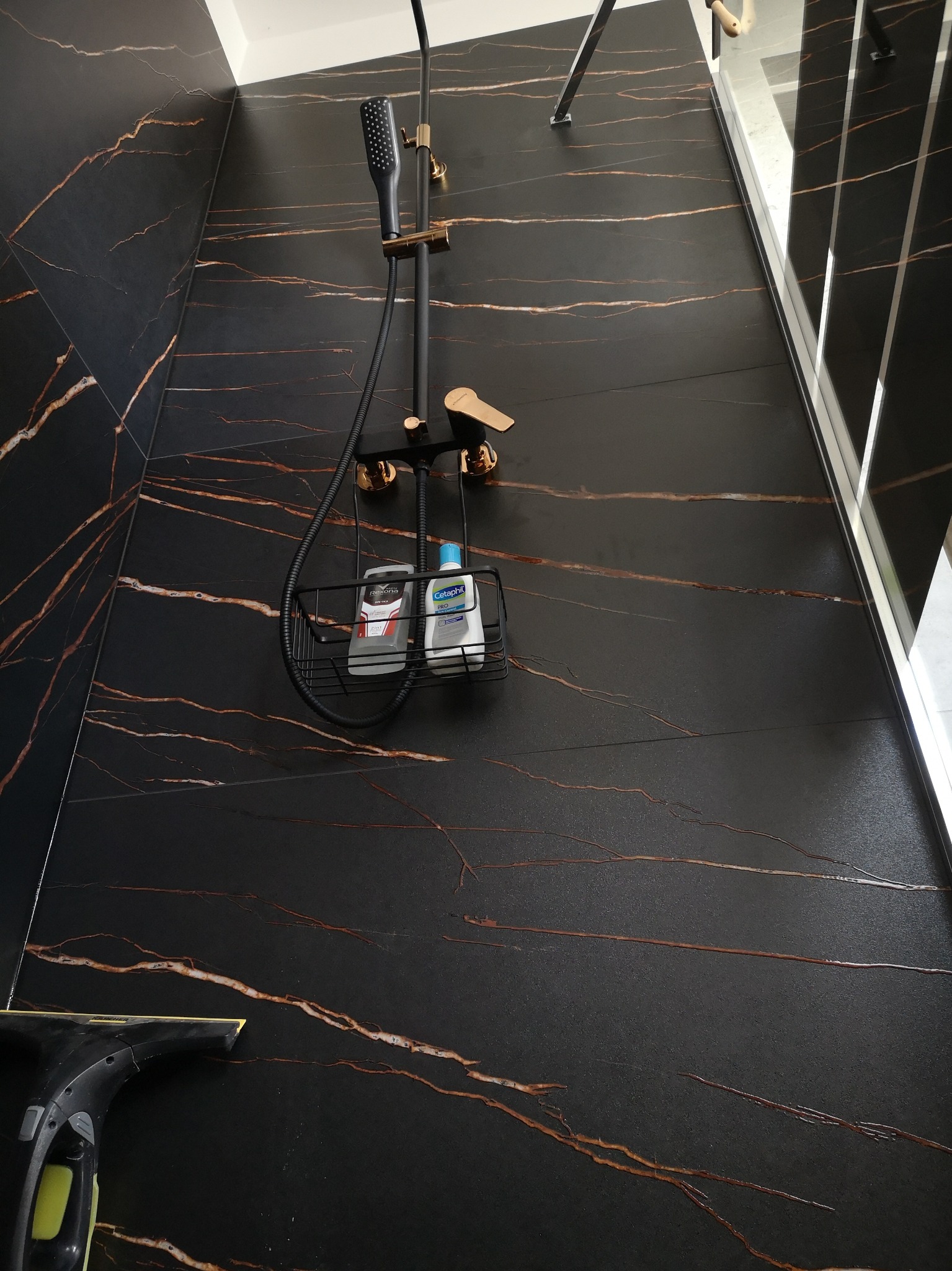 Kabina prysznicowa z czarnymi płytkami imitującymi marmur z brązowymi żyłami, czarna armatura z elementami w kolorze złotym, półka z kosmetykami i myjka do szyb na podłodze.