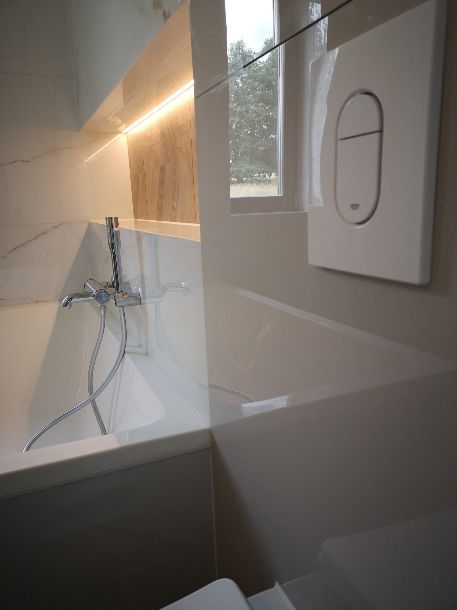 Biała wanna z chromowaną baterią i prysznicem, podświetlana wnęka z drewnopodobnymi płytkami, biały panel spłuczki Grohe na tle okna