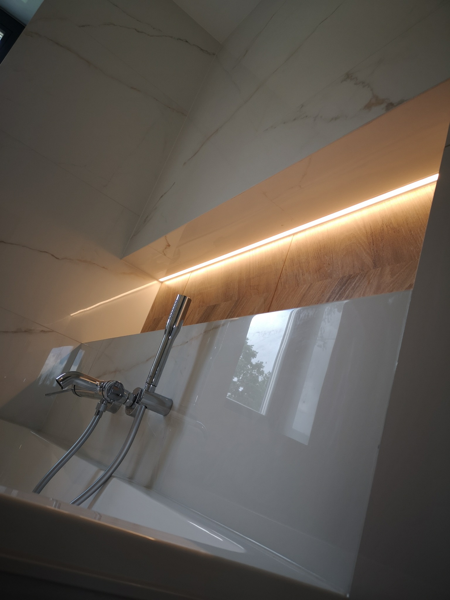 Nowoczesna bateria wannowa z chromowanym wykończeniem na tle białych płytek imitujących marmur i wnęki z podświetleniem LED, odbicie okna w powierzchni wanny.