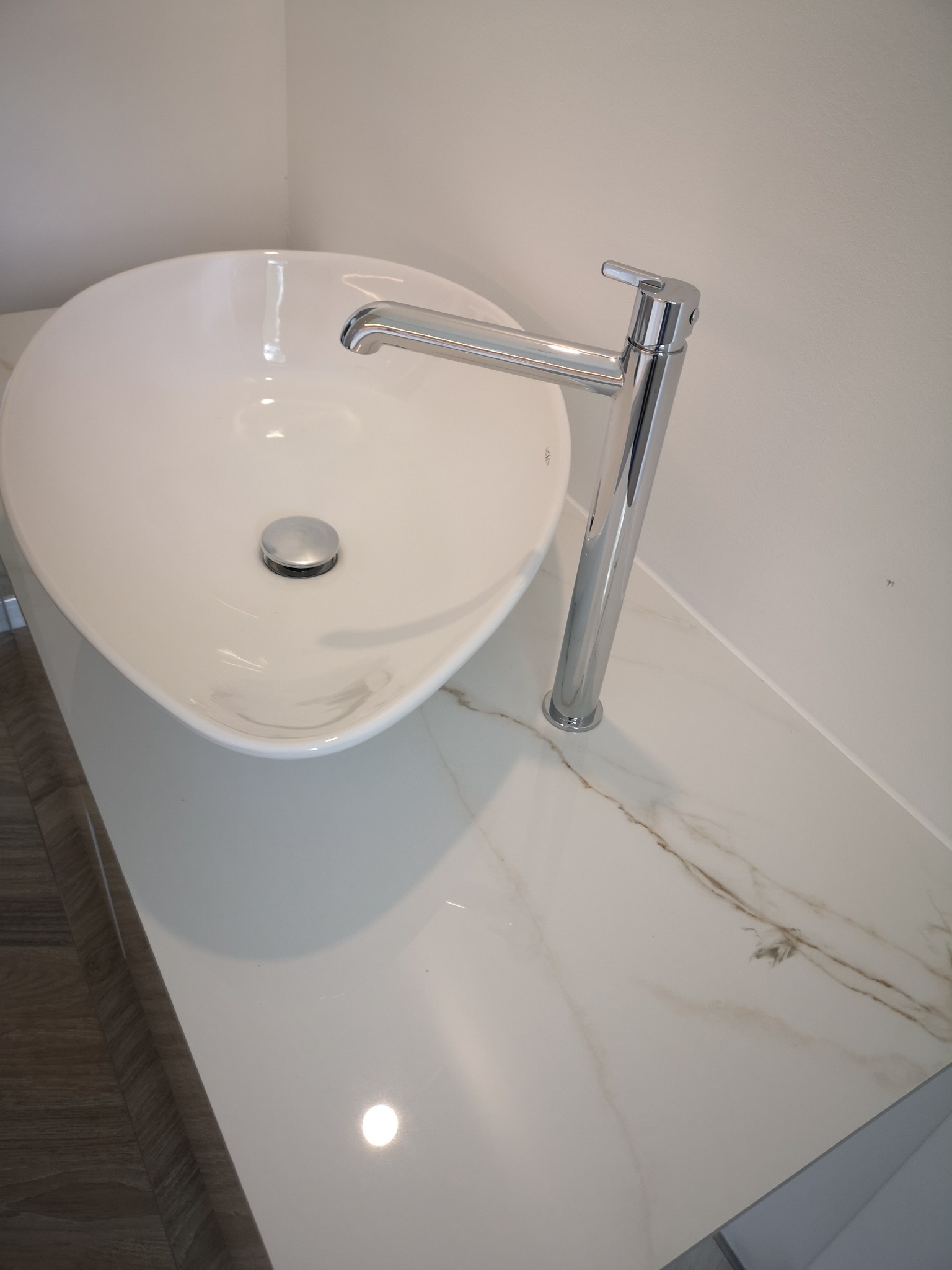 Biała, owalna umywalka nablatowa z chromowaną baterią na blacie z białego marmuru z brązowymi żyłami, zrealizowana w Słupsku.
