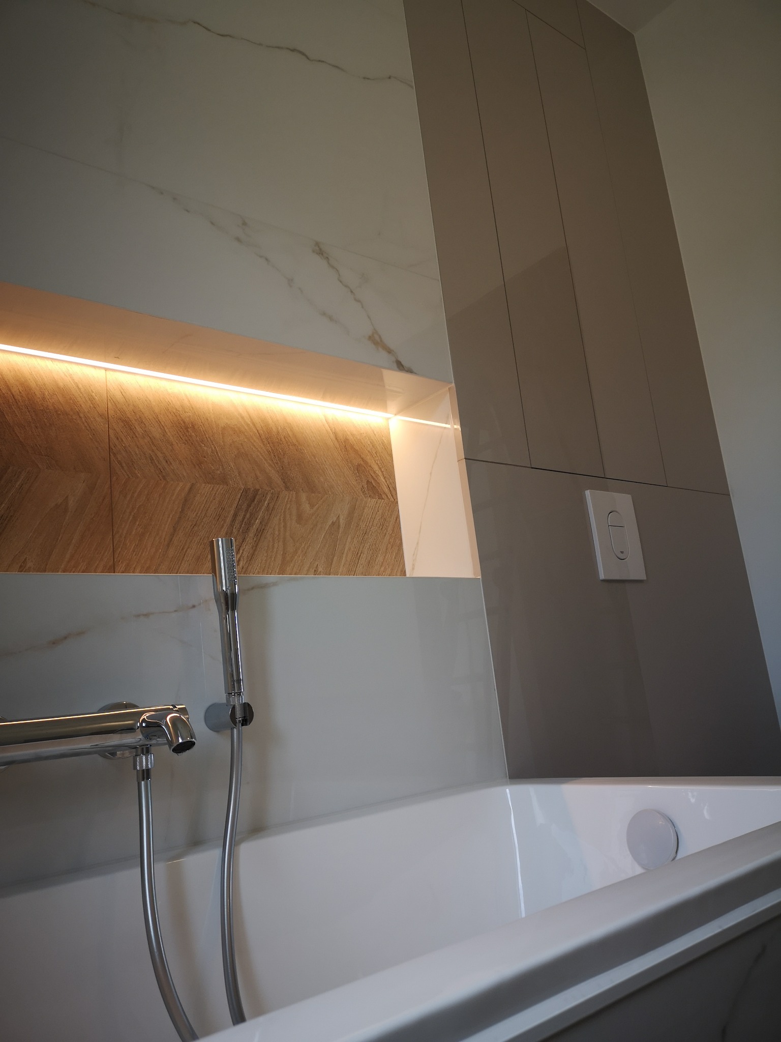 Nowoczesna łazienka z wanną, chromowaną armaturą i podświetlaną wnęką z drewnopodobną okładziną, obok pionowe panele w odcieniach szarości.