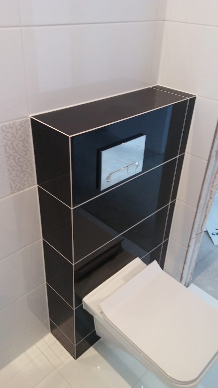 Podwieszana toaleta z czarną obudową spłuczki, wykończona białymi płytkami z delikatnym wzorem, srebrny przycisk spłukiwania.