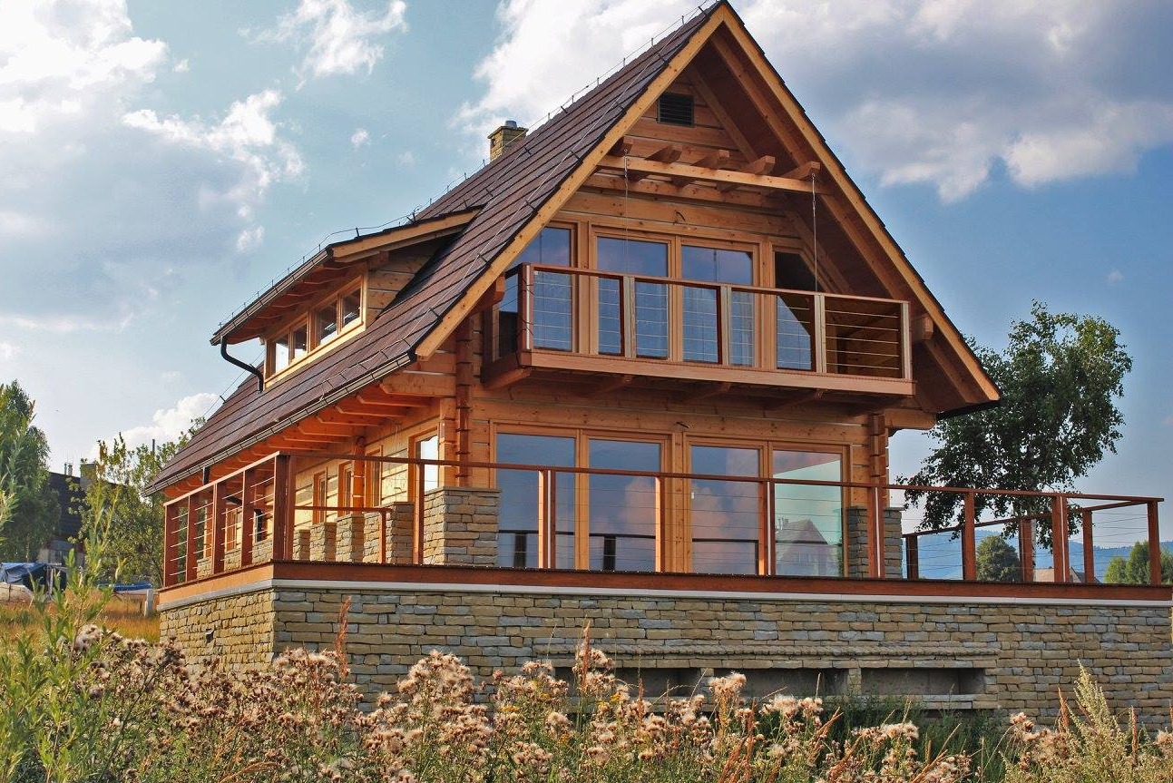Dwukondygnacyjny dom z bali z balkonem na kamiennej podmurówce, brązowy dach, jasne drewno, widok z dołu na tle błękitnego nieba z chmurami.