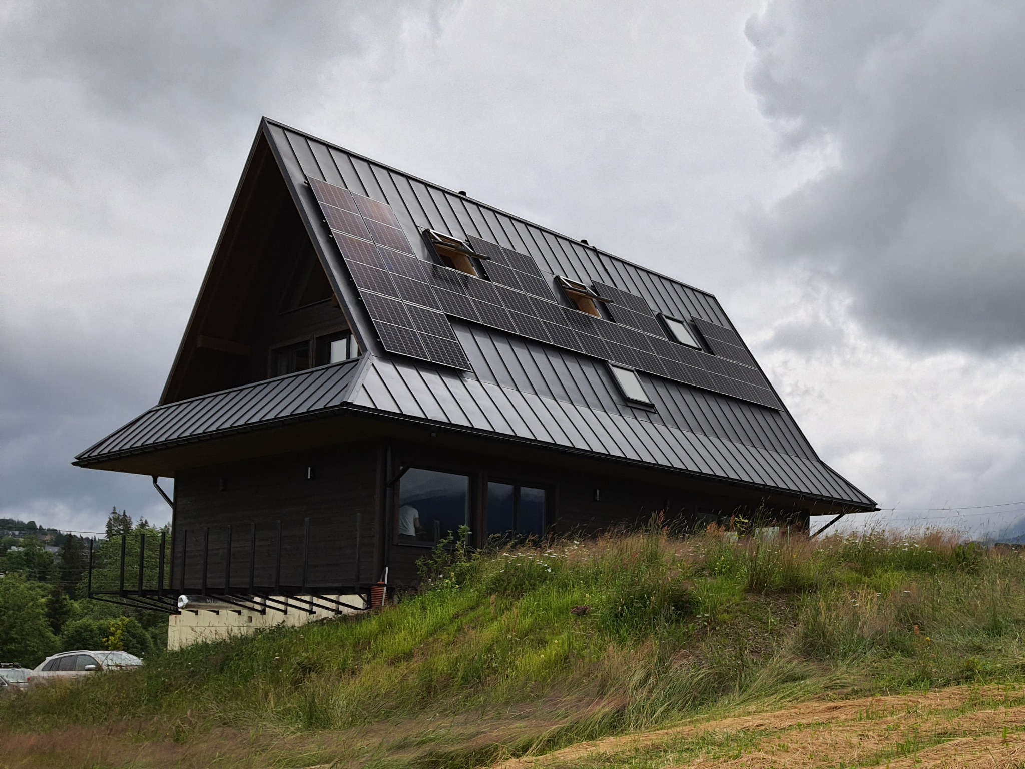 Nowoczesny dom z ciemnego drewna z panelami słonecznymi na dachu, widok z dołu na wzgórzu porośniętym trawą, pochmurne niebo.