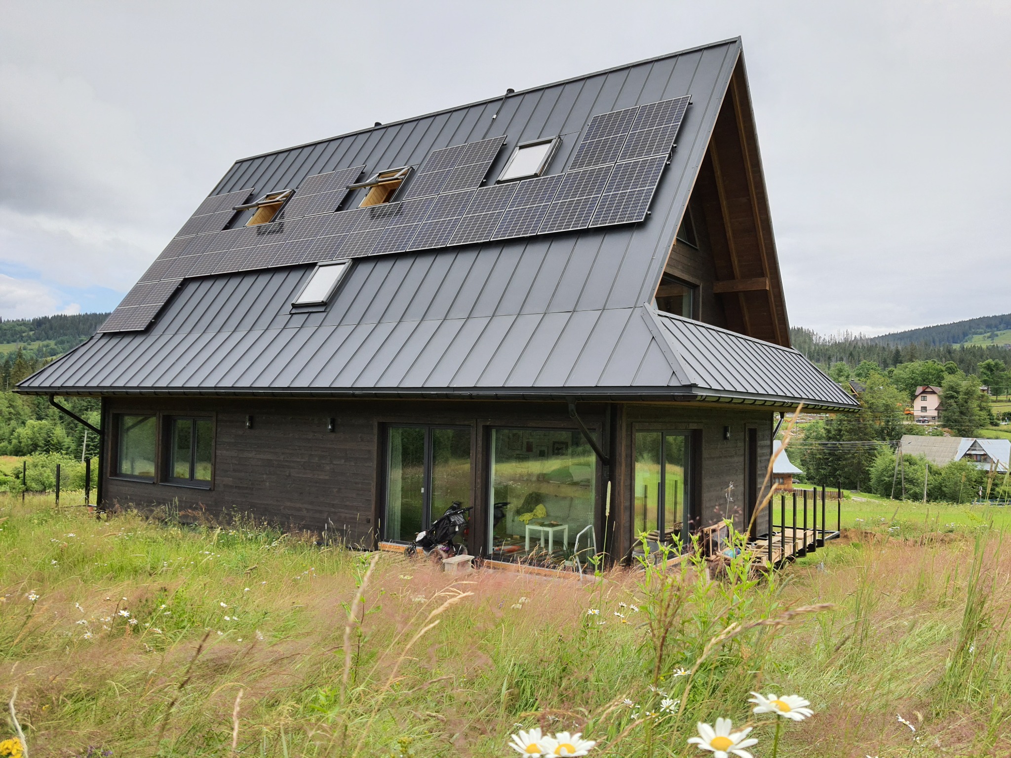 Nowoczesny dom szkieletowy z ciemnego drewna z panelami słonecznymi na dachu, otoczony łąką z kwitnącymi stokrotkami, na tle zalesionych wzgórz.
