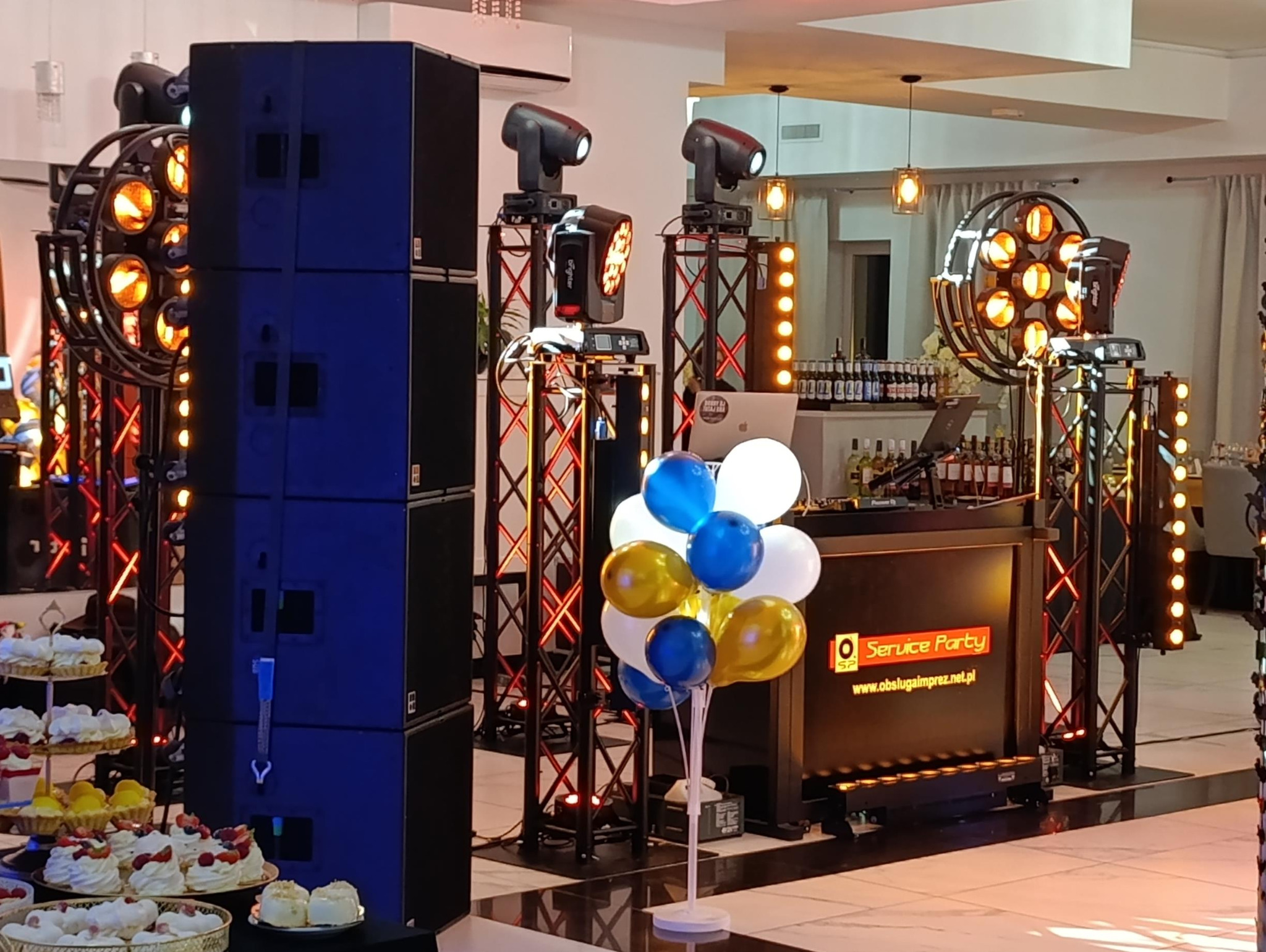 Scena z nagłośnieniem, oświetleniem i stanowiskiem DJ na imprezie. Widoczne kolumny głośnikowe, reflektory, dekoracje balonowe i słodki stół. Logo firmy na stanowisku DJ.