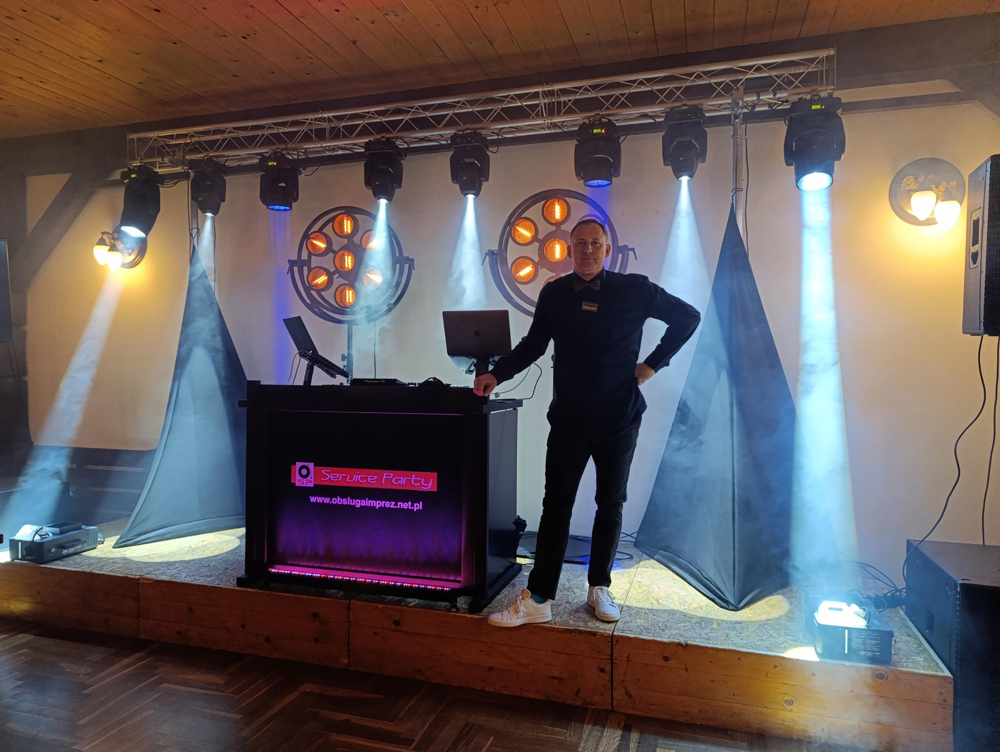 DJ na podwyższeniu, z oświetleniem scenicznym i dekoracjami. Widoczny sprzęt DJ-ski z logo 'Service Party'. Scena oświetlona reflektorami, tworząc klimat imprezy.