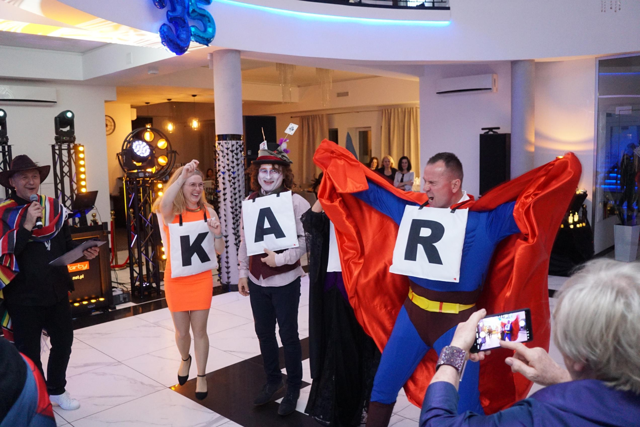 Uczestnicy imprezy w kostiumach z literami 'K', 'A', 'R', tworzący napis 'KAR' podczas występu na scenie. W tle oświetlenie sceniczne i publiczność.