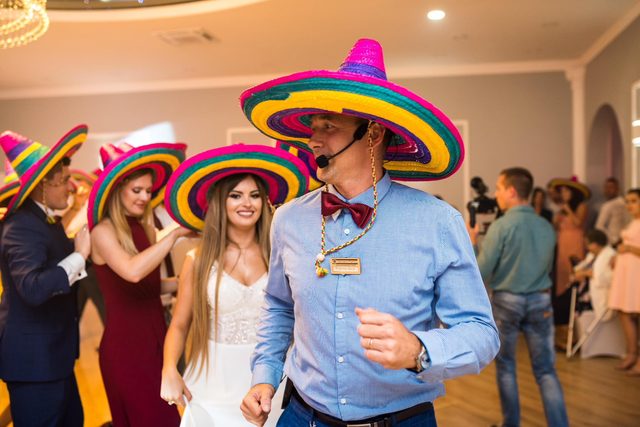 Uśmiechnięty wodzirej w kolorowym sombrero i muszce prowadzi zabawę taneczną z gośćmi weselnymi, również w sombrerach. W tle sala balowa.