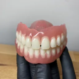 Górna proteza zębową, trzymana w dłoniach w czarnych rękawiczkach, prezentowana na drewnianym blacie na tle szarej ściany.