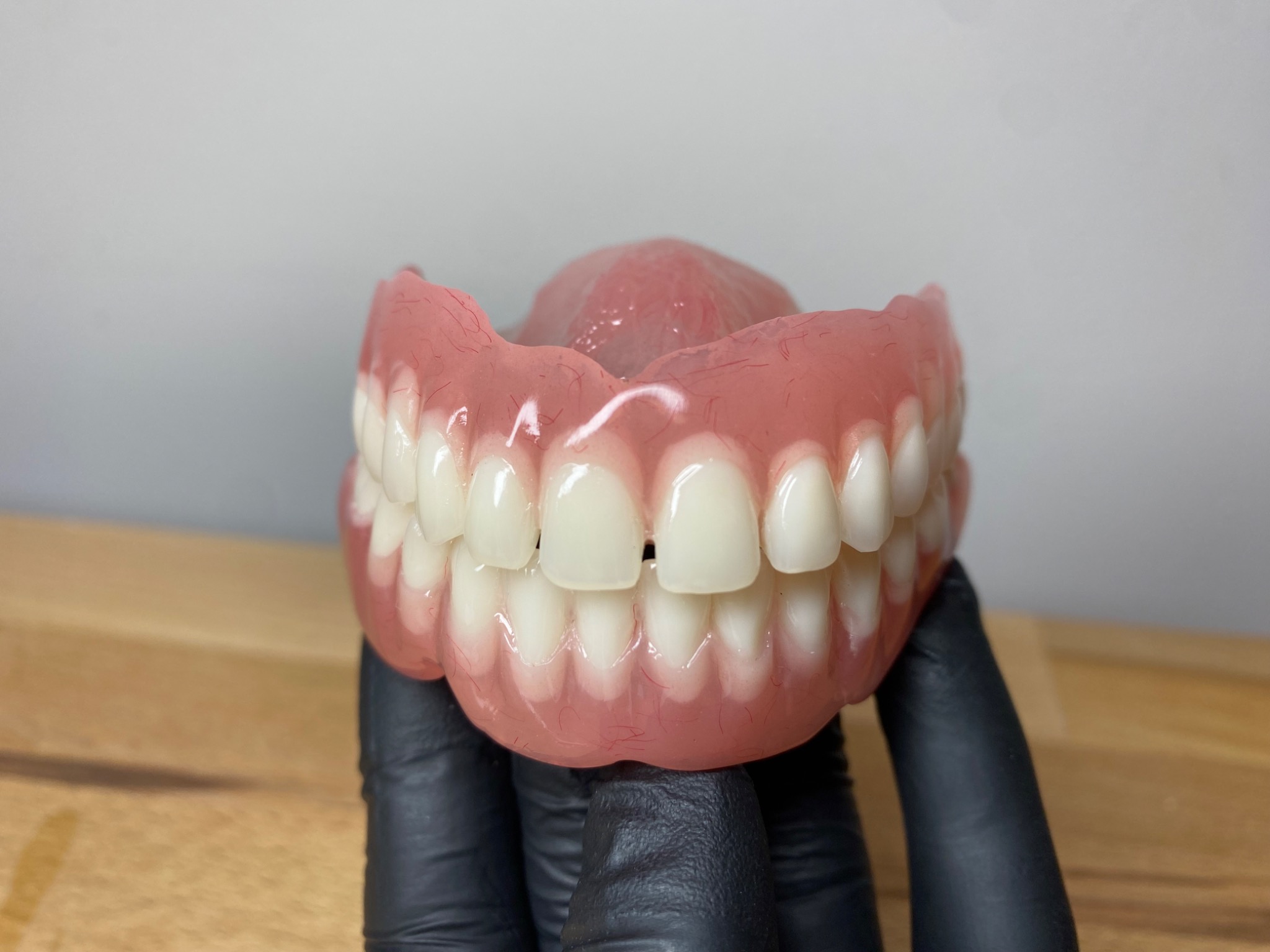 Górna proteza zębową, trzymana w dłoniach w czarnych rękawiczkach, prezentowana na drewnianym blacie na tle szarej ściany.